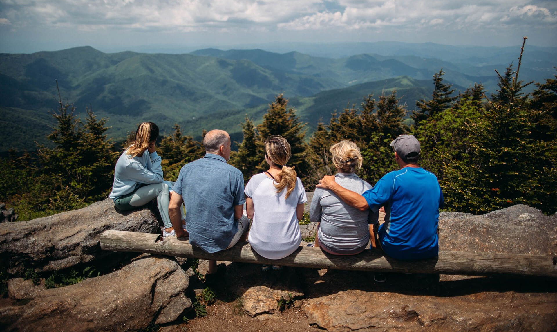 Five people seated on a log bench, facing a mountain vista on a sunny day.
