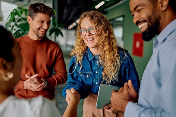 Group of people smiling, shaking hands in office setting. Person in denim shirt, holding tablet, shakes hands with another. Group of people smiling, shaking hands in office setting. Person in denim shirt, holding tablet, shakes hands with another.