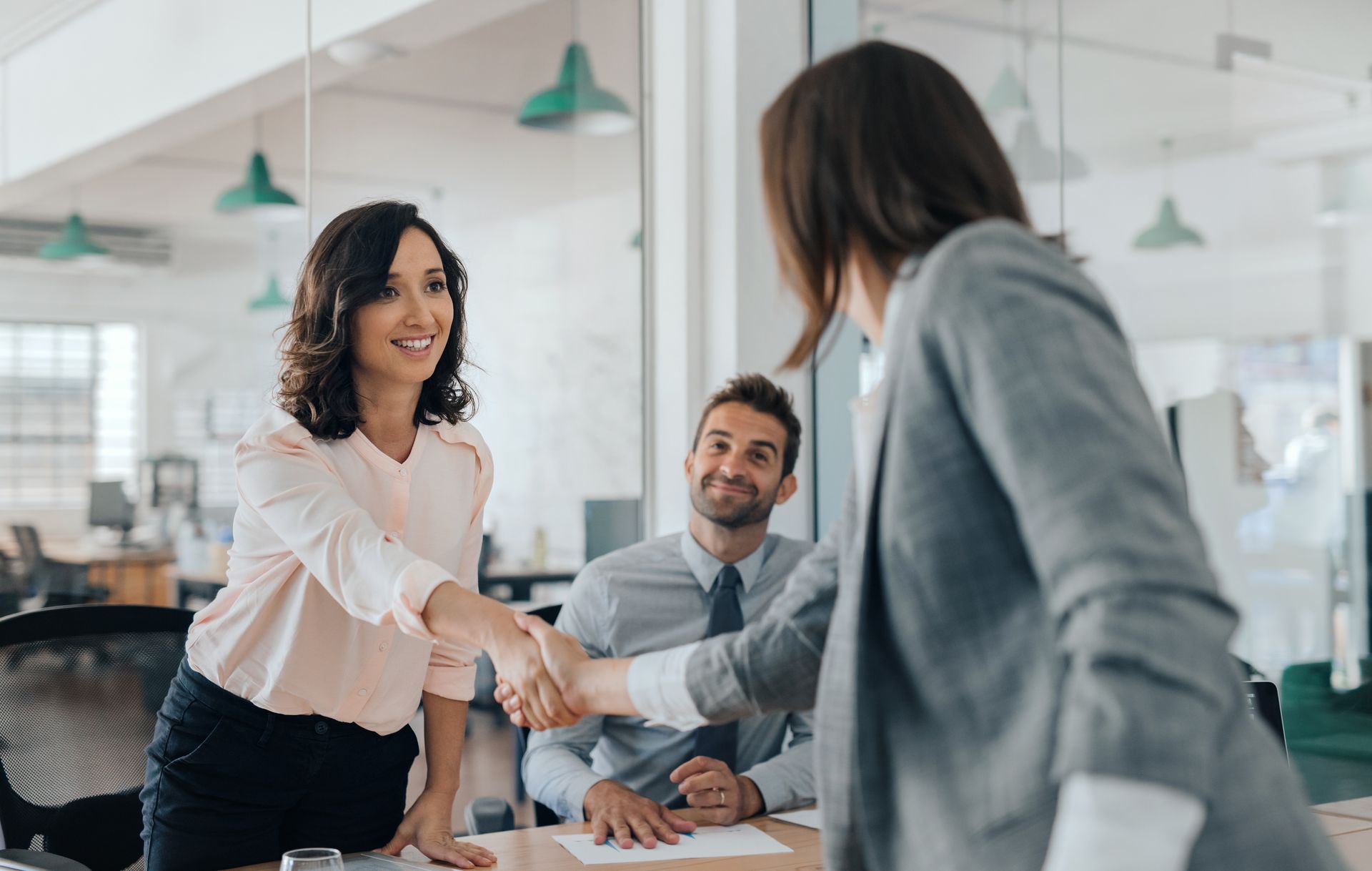 Woman shaking hands with another woman in a bright office; a man smiles in the background. Woman shaking hands with another woman in a bright office; a man smiles in the background.