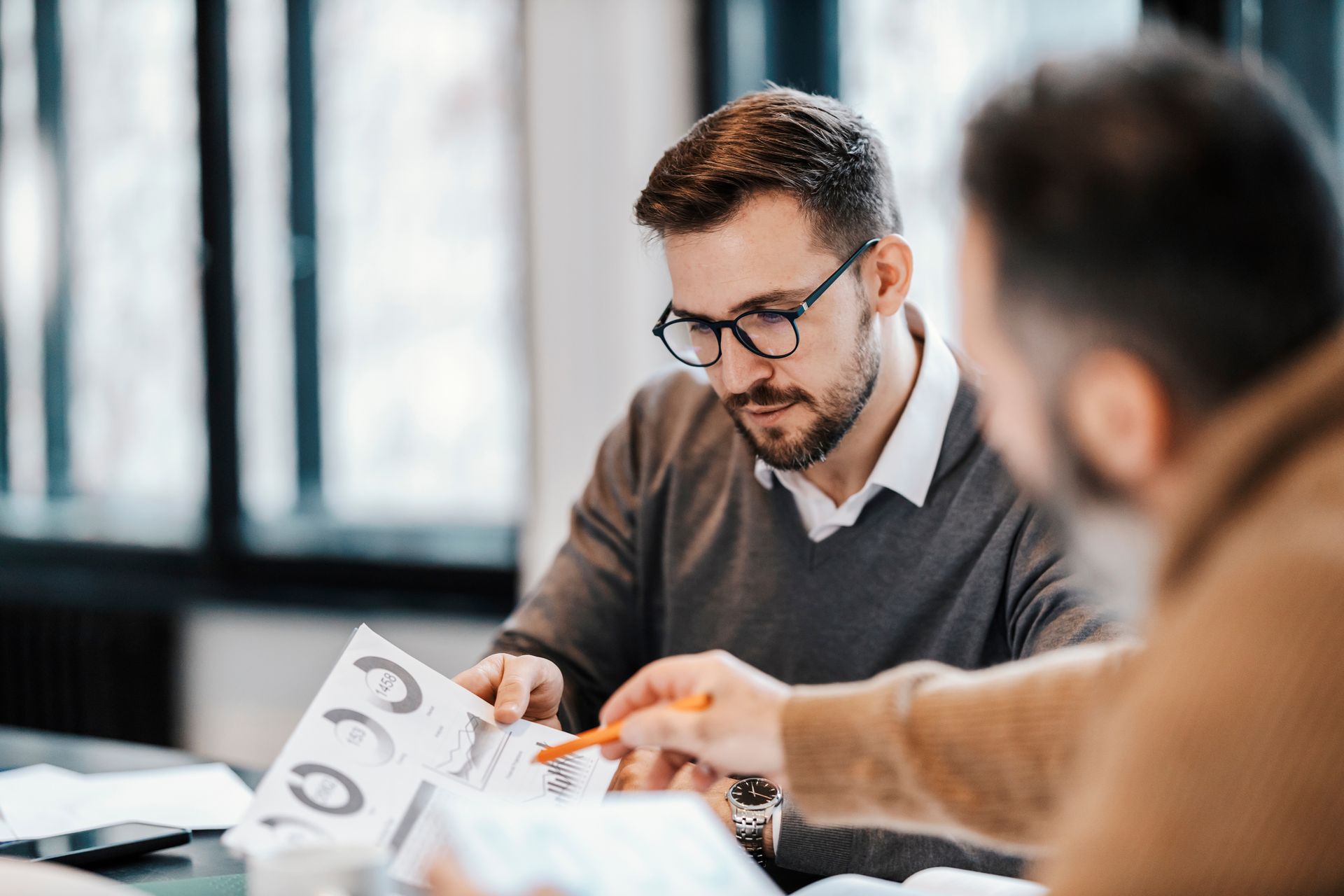 Man with glasses looks at document, other man points at it. Both are in an office setting. Man with glasses looks at document, other man points at it. Both are in an office setting.