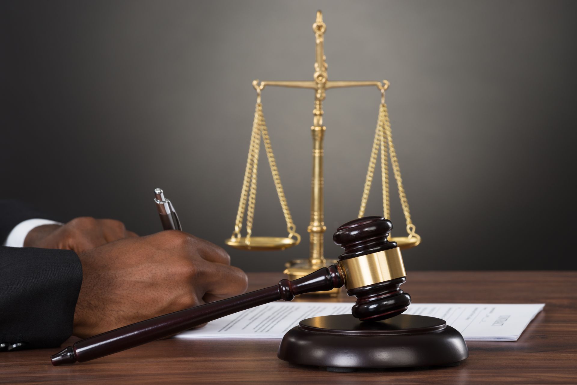 An attorney’s hands writing on a document at a wooden desk with the scales of justice and gavel. An attorney’s hands writing on a document at a wooden desk with the scales of justice and gavel.