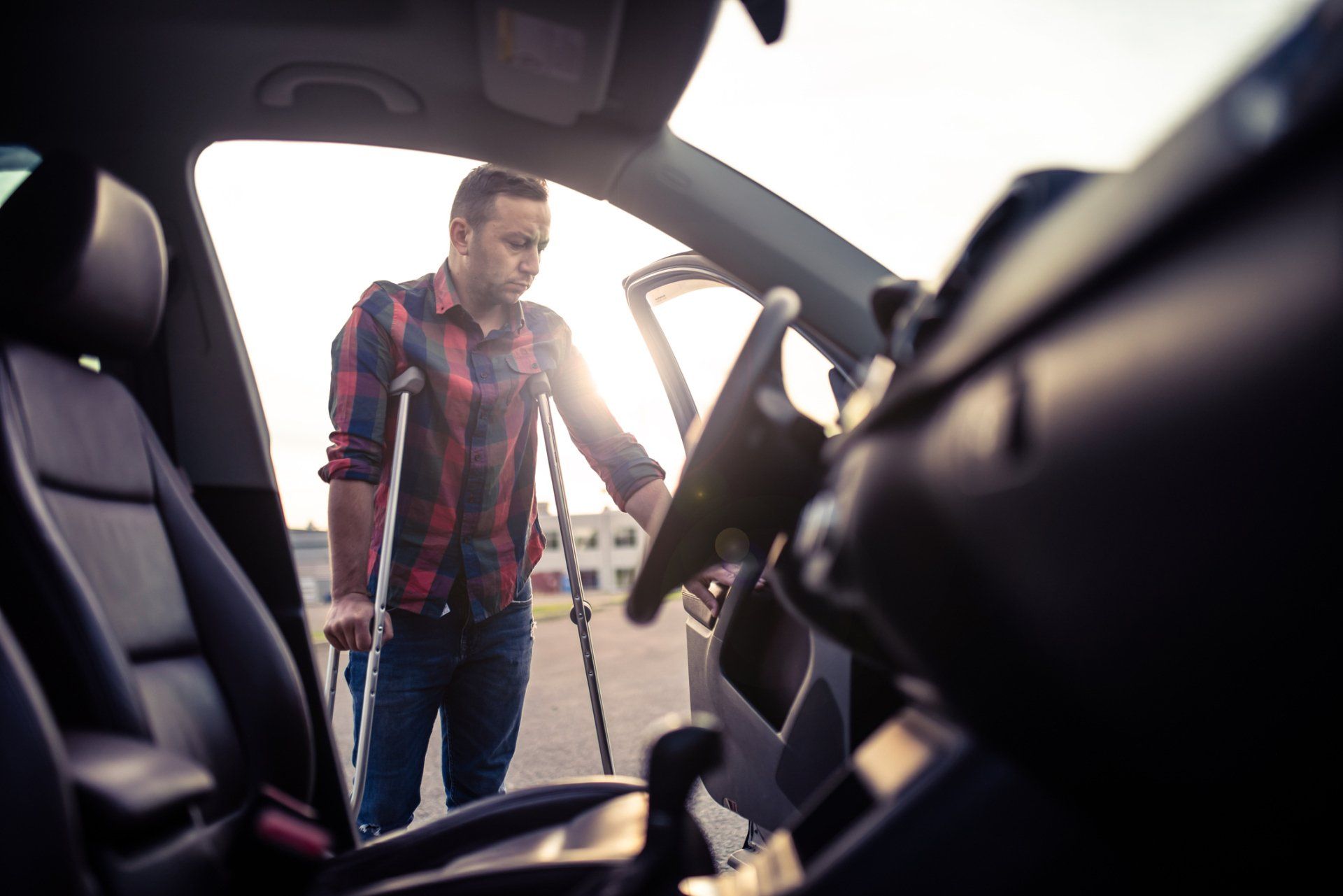Man with crutches getting into a car, wearing plaid shirt and jeans. Bright sunlight in the background.