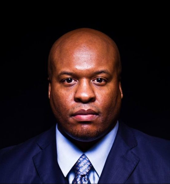 Attorney Theodis | Little Rock, AR | Thompson Law Firm, PLLC Bald Black man in a suit, serious expression, dark background.