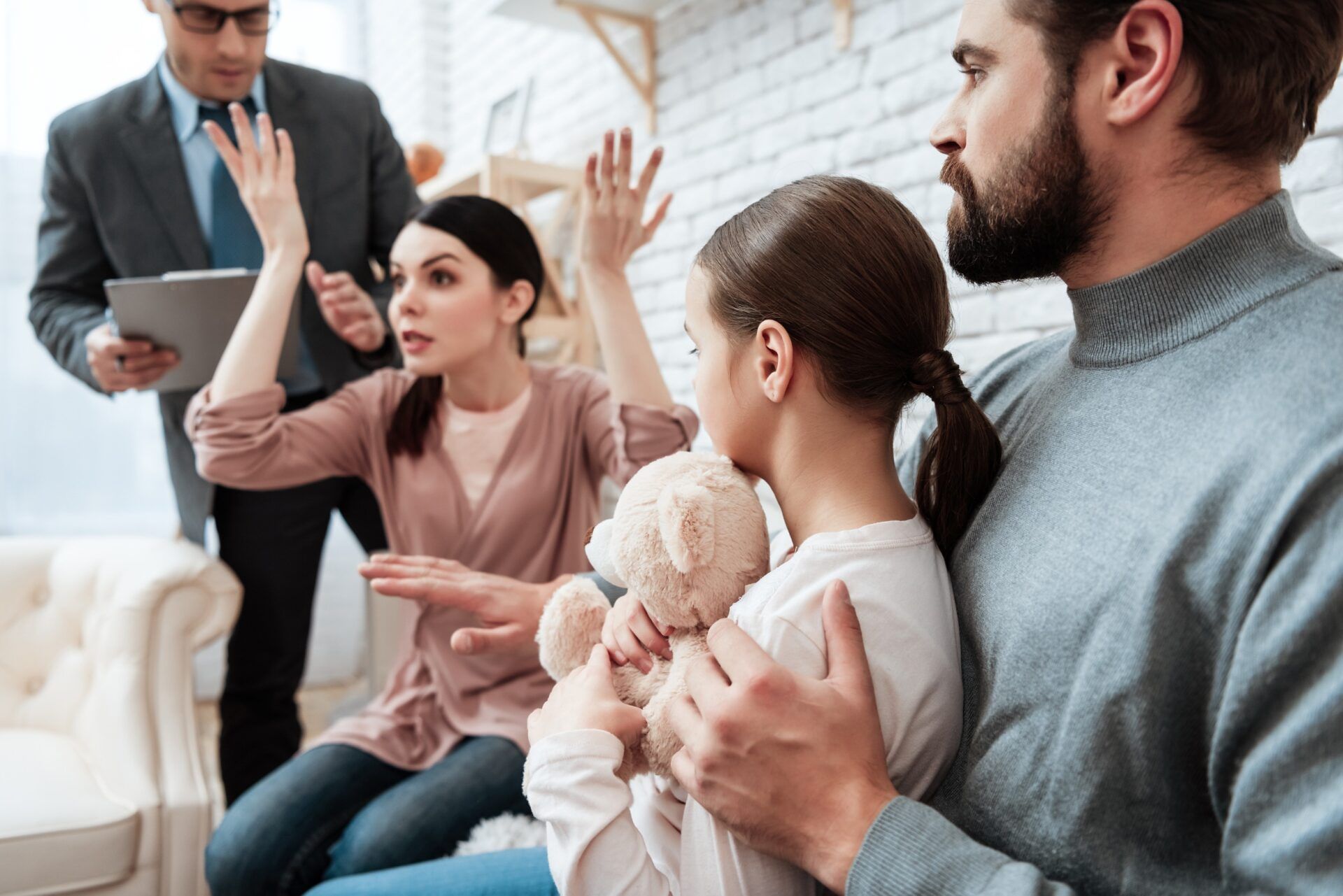 a family is sitting on a couch with a teddy bear while a man and woman argue .