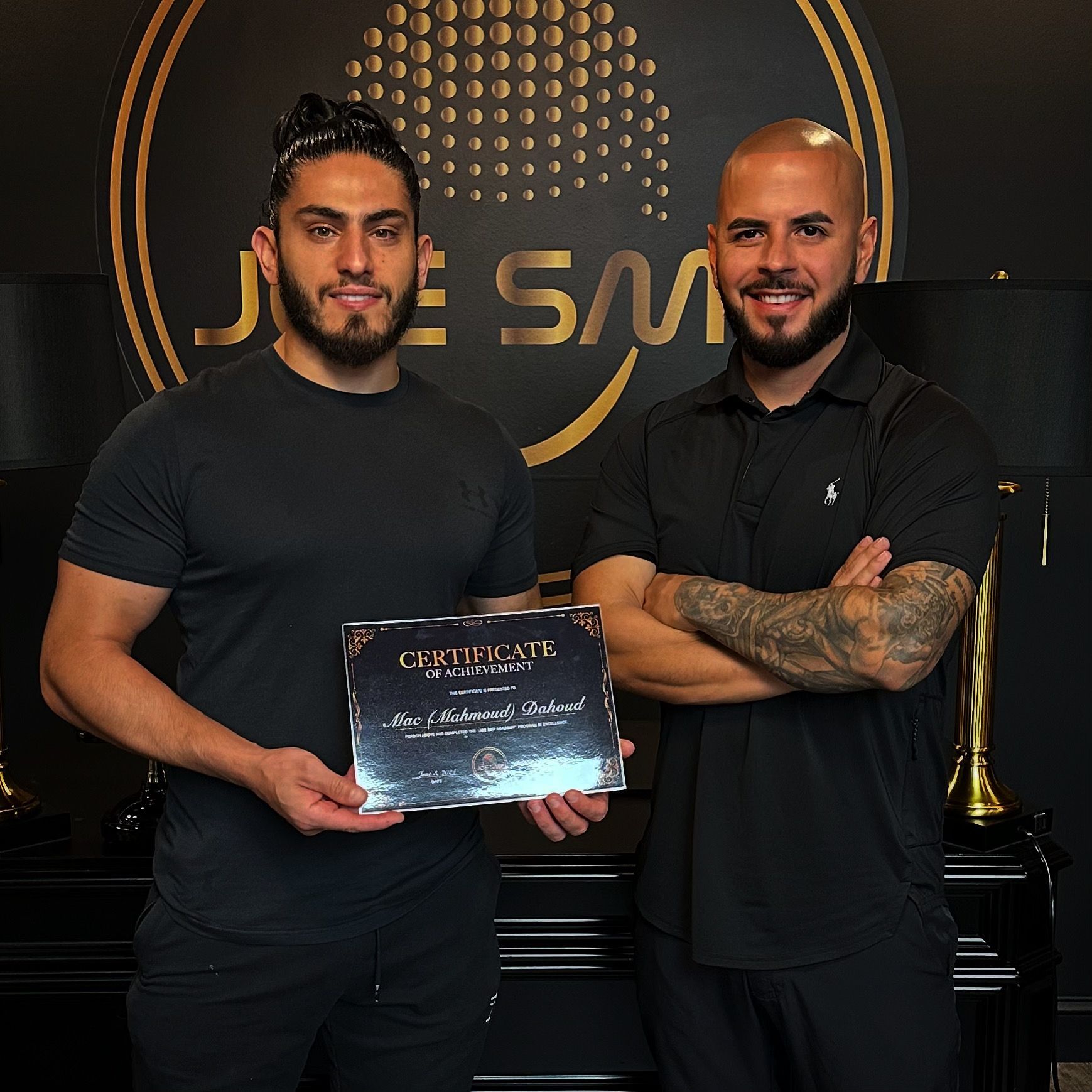 Two men in a black room. One holds a certificate, the other has arms crossed. A logo is in the background.