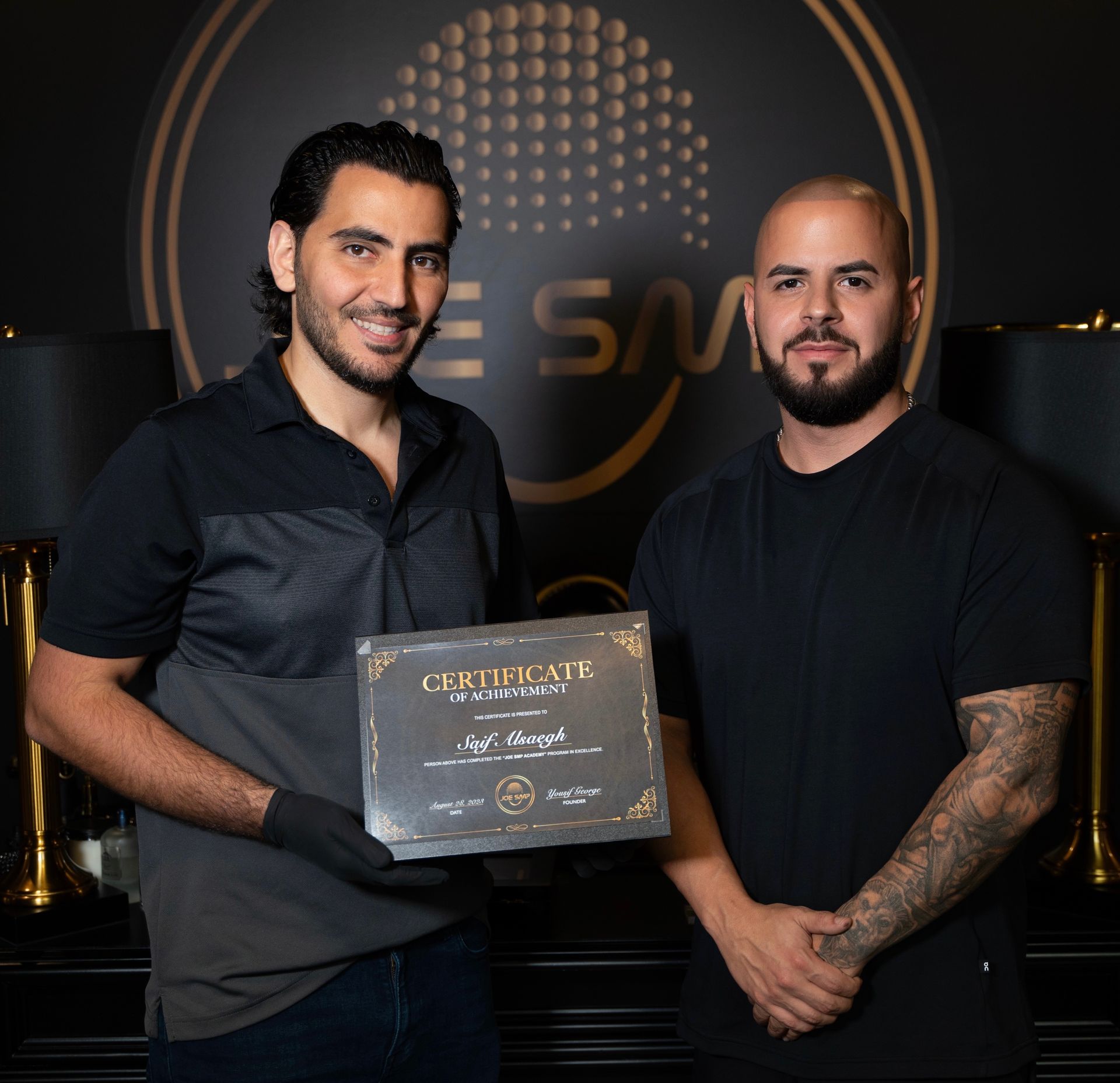Two men holding a certificate in front of a logo. The man on the left smiles, holding the certificate. The man on the right smiles.