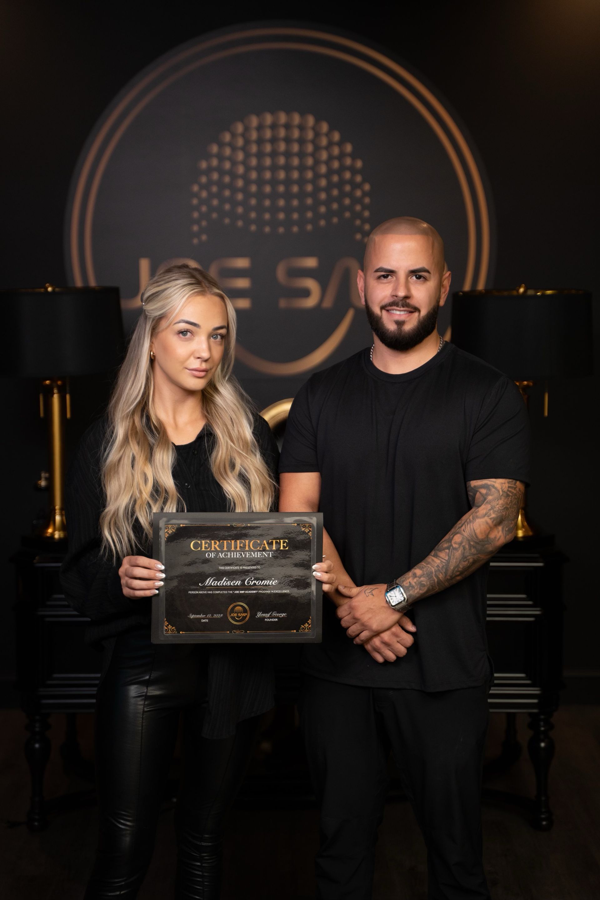 Woman and man hold a certificate, smiling.  Dark background with logo, possibly in a studio setting.