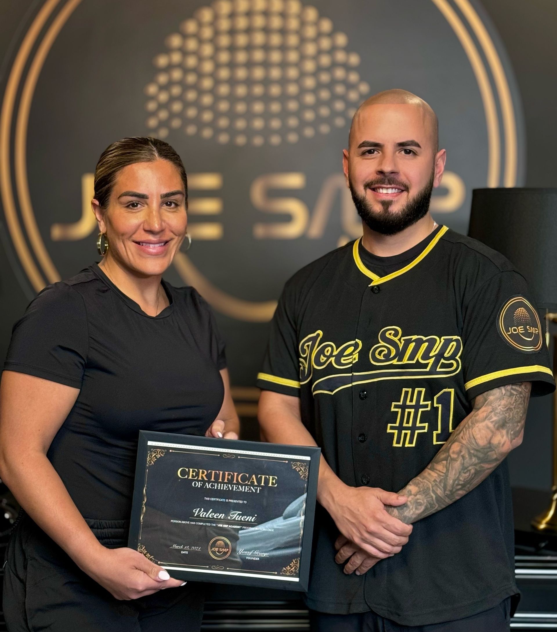 Woman and man holding certificate; Joe Sap logo in background. Man in baseball jersey smiles.
