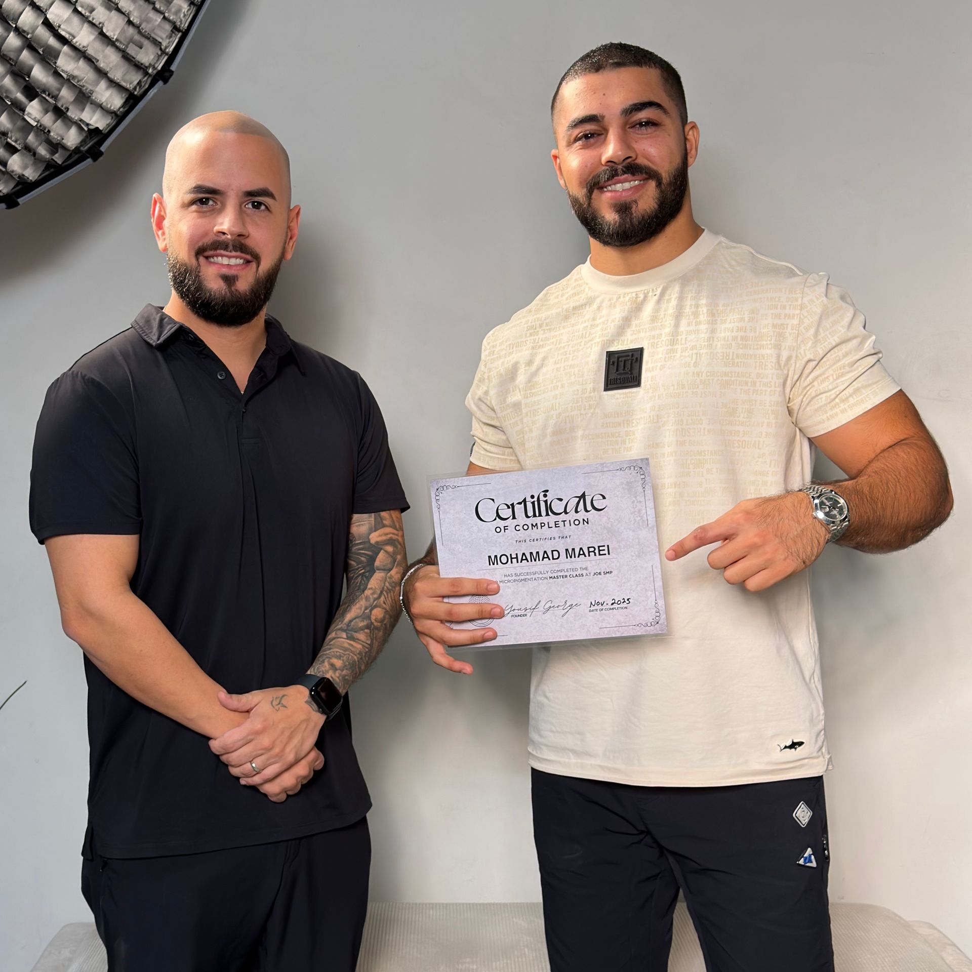 Two men hold a certificate, one pointing. They stand in front of a white wall, smiling.
