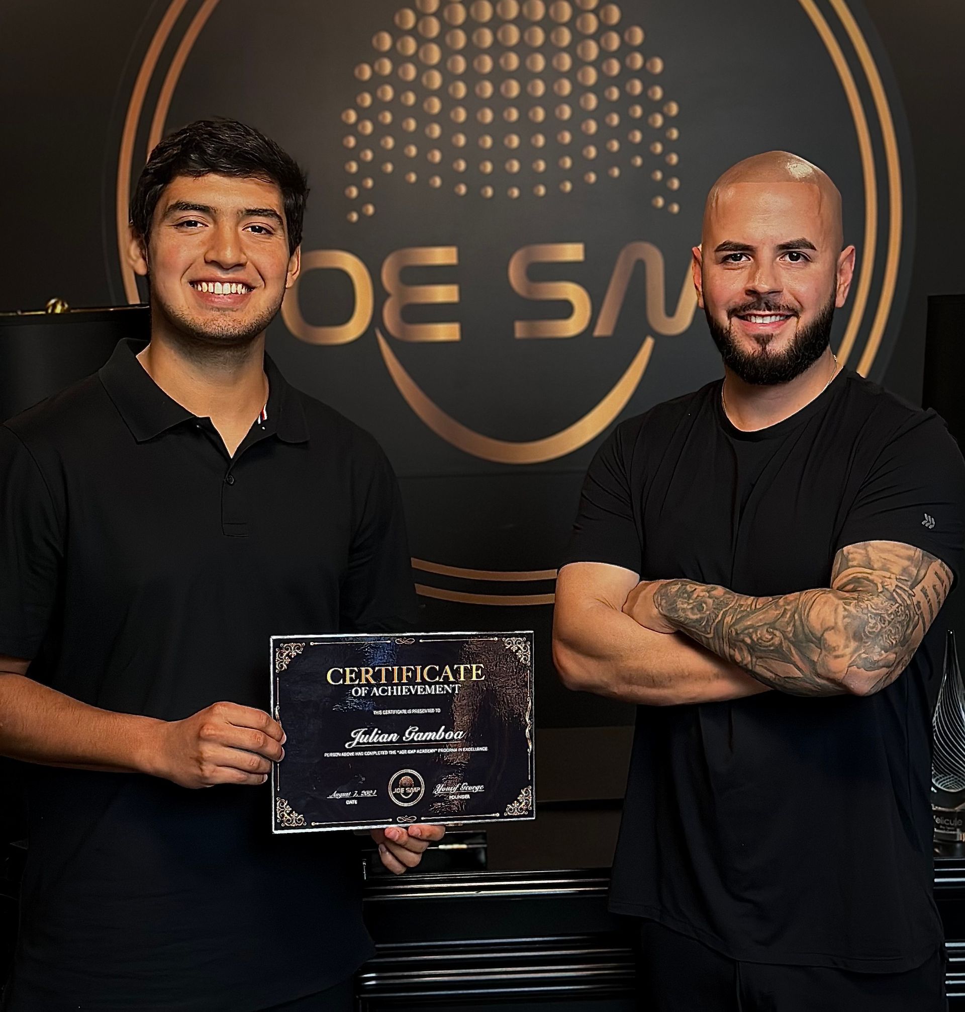Two men smiling, one holding a certificate. They are in front of a logo that says