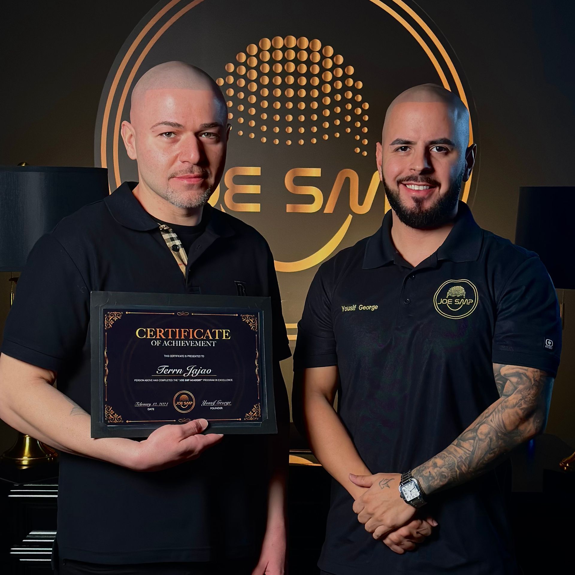 Two men holding a framed certificate in front of a gold logo. They are smiling. One wears a black polo shirt, other has a black shirt.