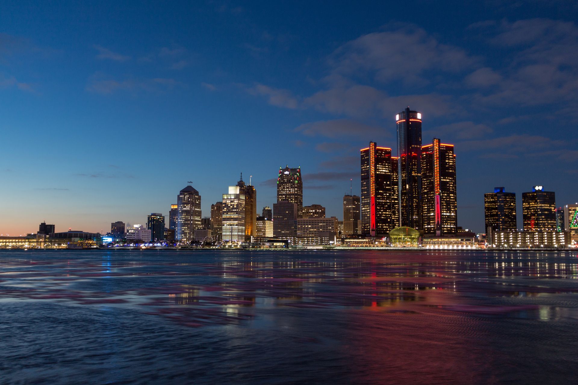 Detroit skyline at dusk reflected in water. Buildings lit with lights against a blue sky.