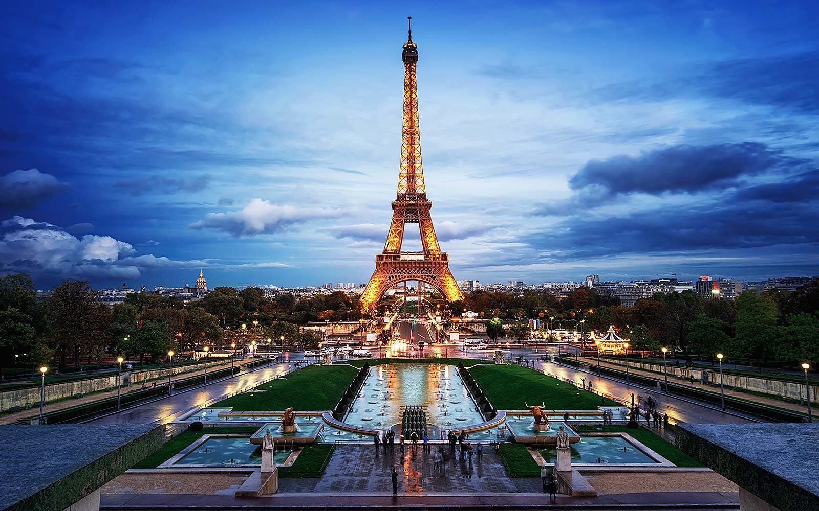 Eiffel Tower lit up at dusk, reflected in a long pool with tiered gardens in Paris.