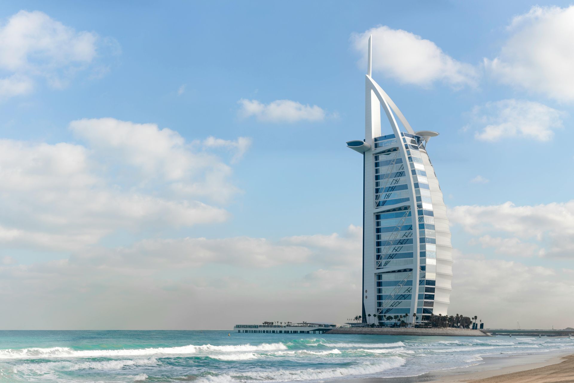 Burj Al Arab hotel in Dubai, a sail-shaped luxury building by the turquoise sea under a cloudy sky.