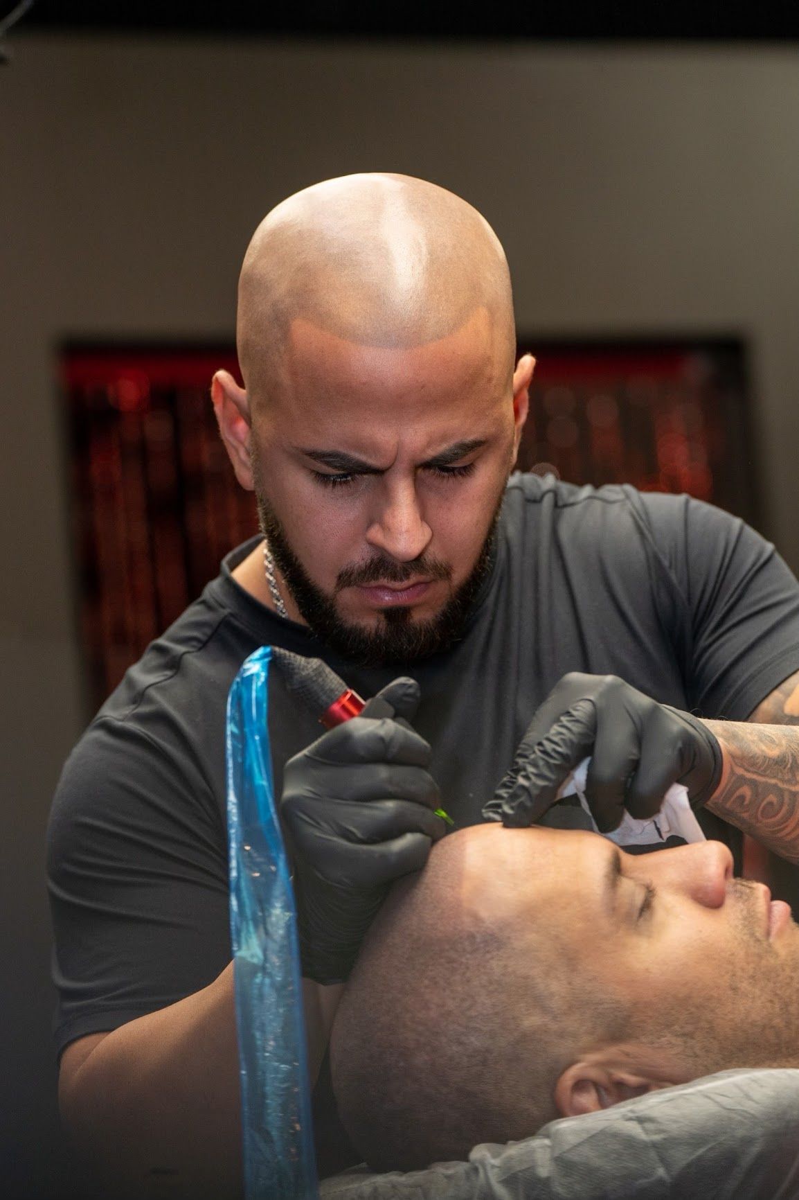 Man tattooing another man's scalp, both with shaved heads, in a studio setting.