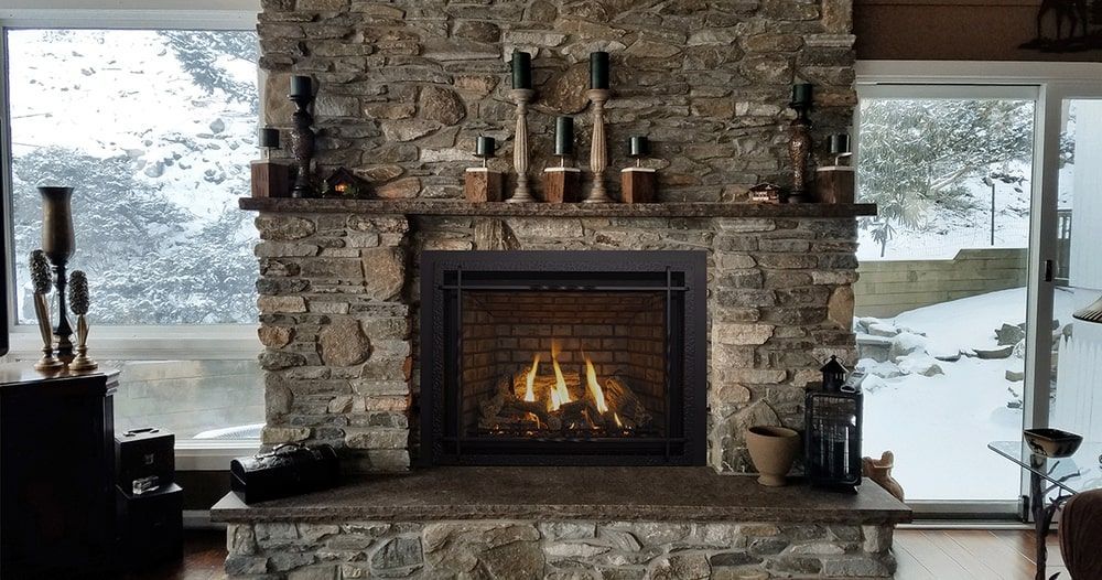 Fireplace with stone surround and lit fire, flanked by large windows with snowy outdoor view, candles on mantel.
