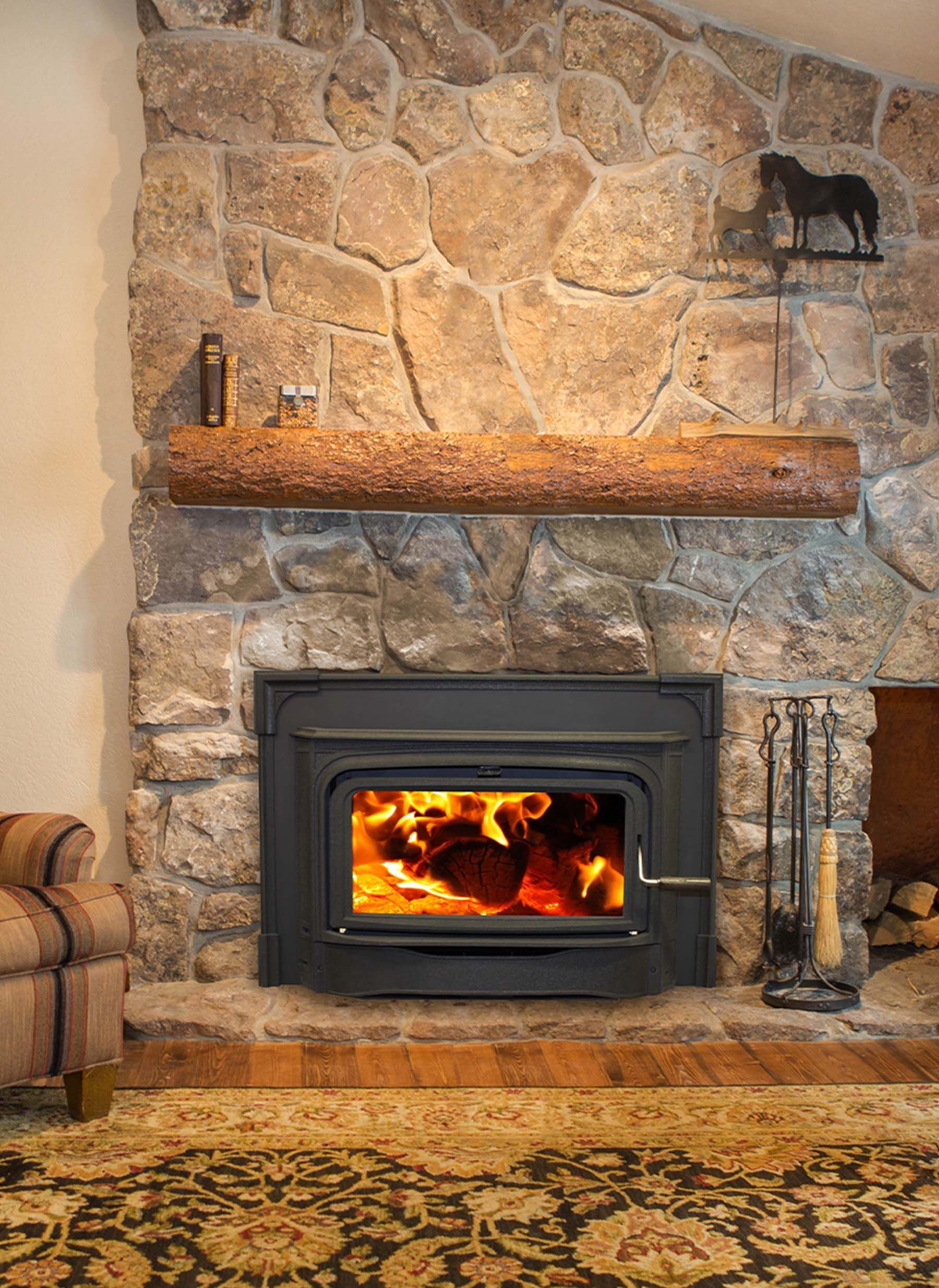 A fireplace with a roaring fire built into a stone wall, topped with a wood beam mantel. A horse weather vane adorns the wall.