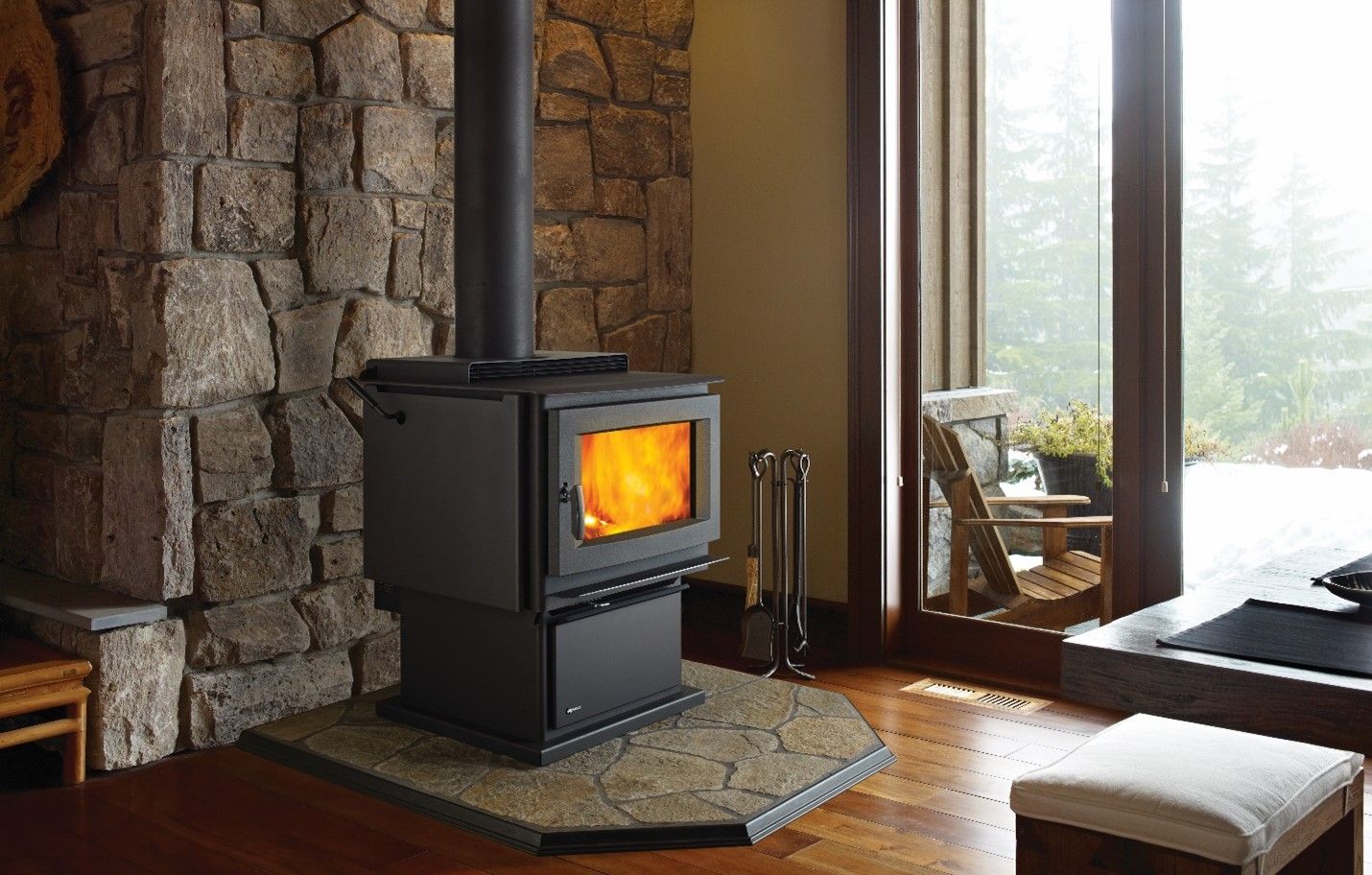 Wood-burning stove in a cozy living room setting with stone wall backdrop and window.