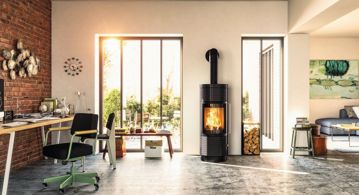 Modern living room with a wood-burning stove centered between large windows. A desk and exposed brick wall are on the left.