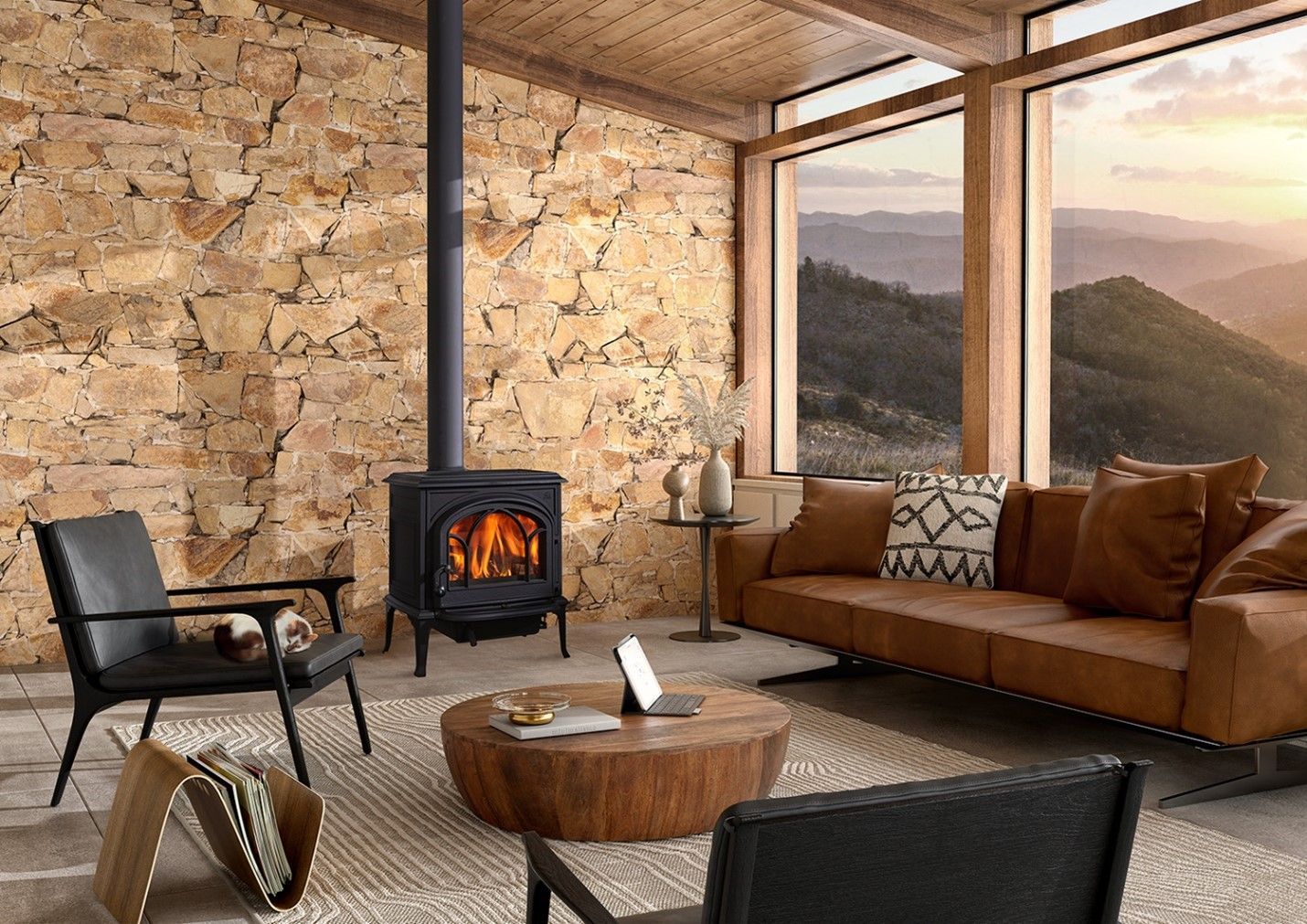 Cozy living room with a lit fireplace, brown leather sofa, and a stone wall. Large windows overlook a mountain view.