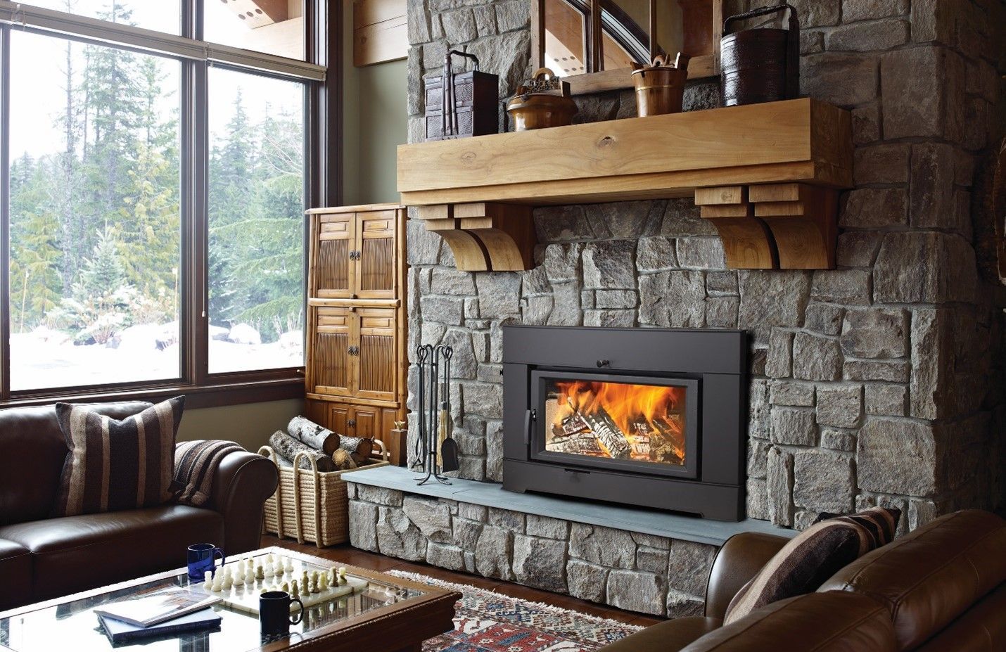 Cozy living room with a stone fireplace, a burning fire, and a wooden mantel. Includes a large window with a snowy outdoor view and leather sofas.