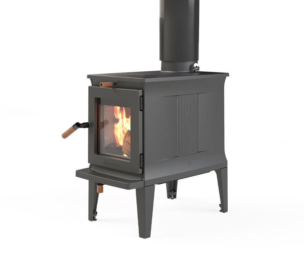 A black wood-burning stove with a lit fire sits on a stone hearth in a log cabin, with a large window overlooking a wooded area.