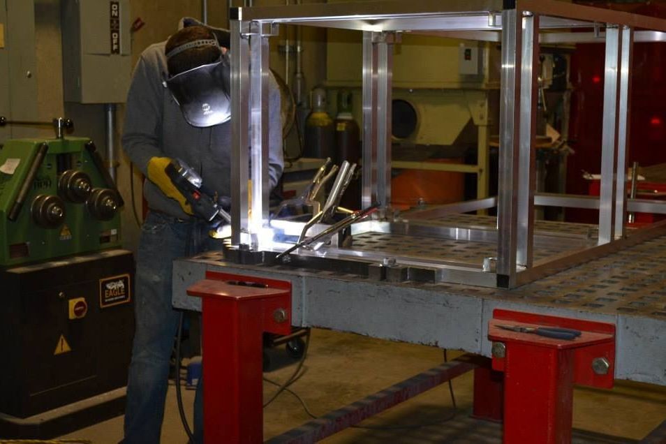 A welder in a shop welds a metal frame, wearing a helmet and gloves. The setting has other machinery and red supporting structures.