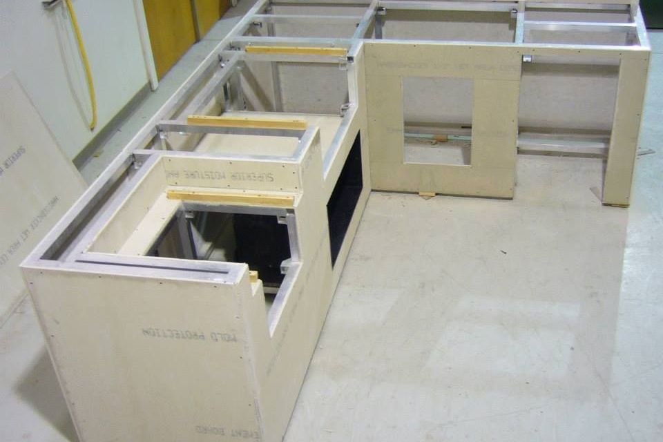 Partially constructed kitchen cabinets with a metal frame and light-colored panels, set on a light-colored floor.