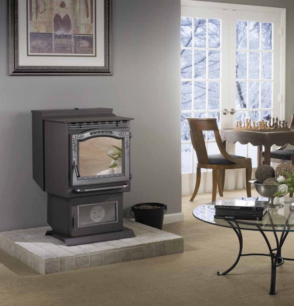 A dark gray pellet stove on a light gray tile hearth in a living room. Windows and a table are in the background.