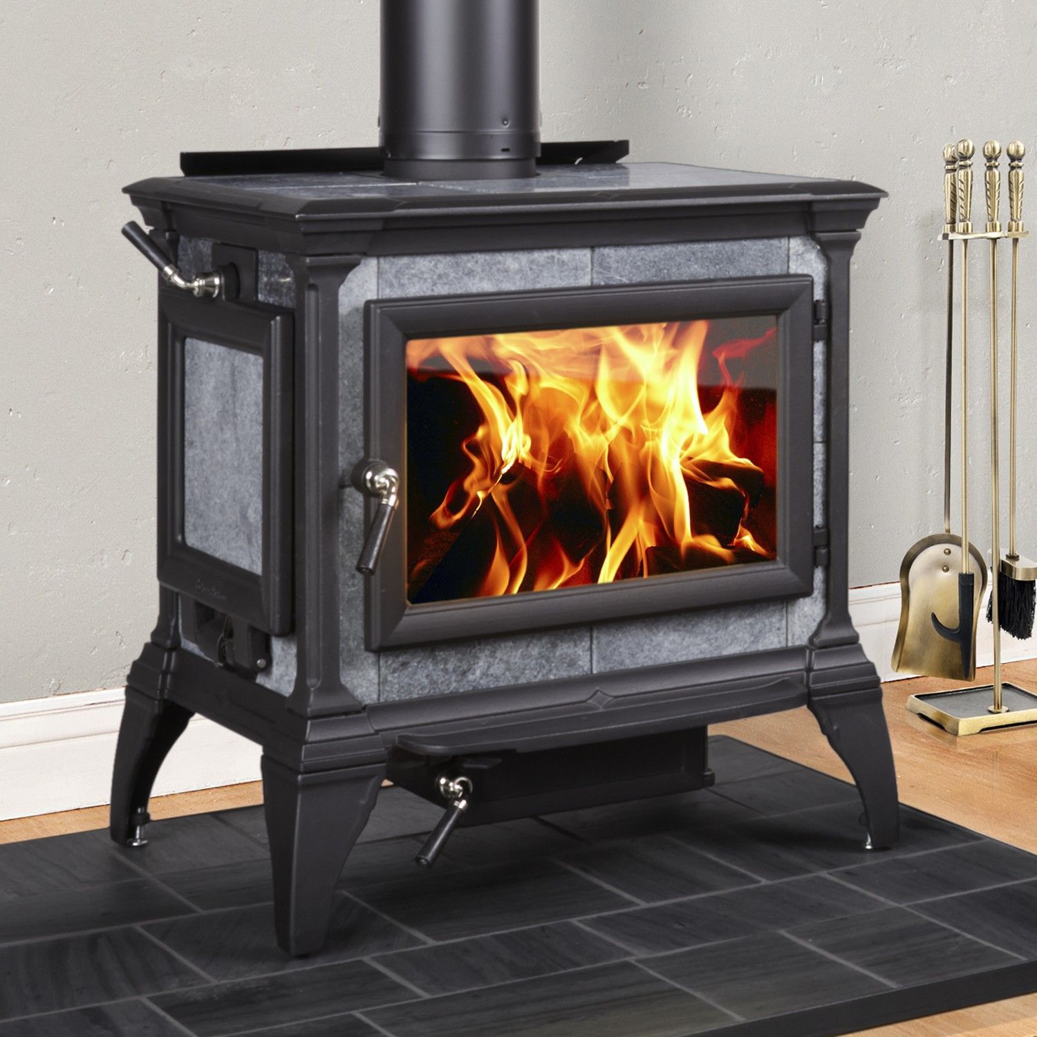 A wood-burning stove with flames visible through a glass door, set on a tiled hearth. Accessories stand beside it.