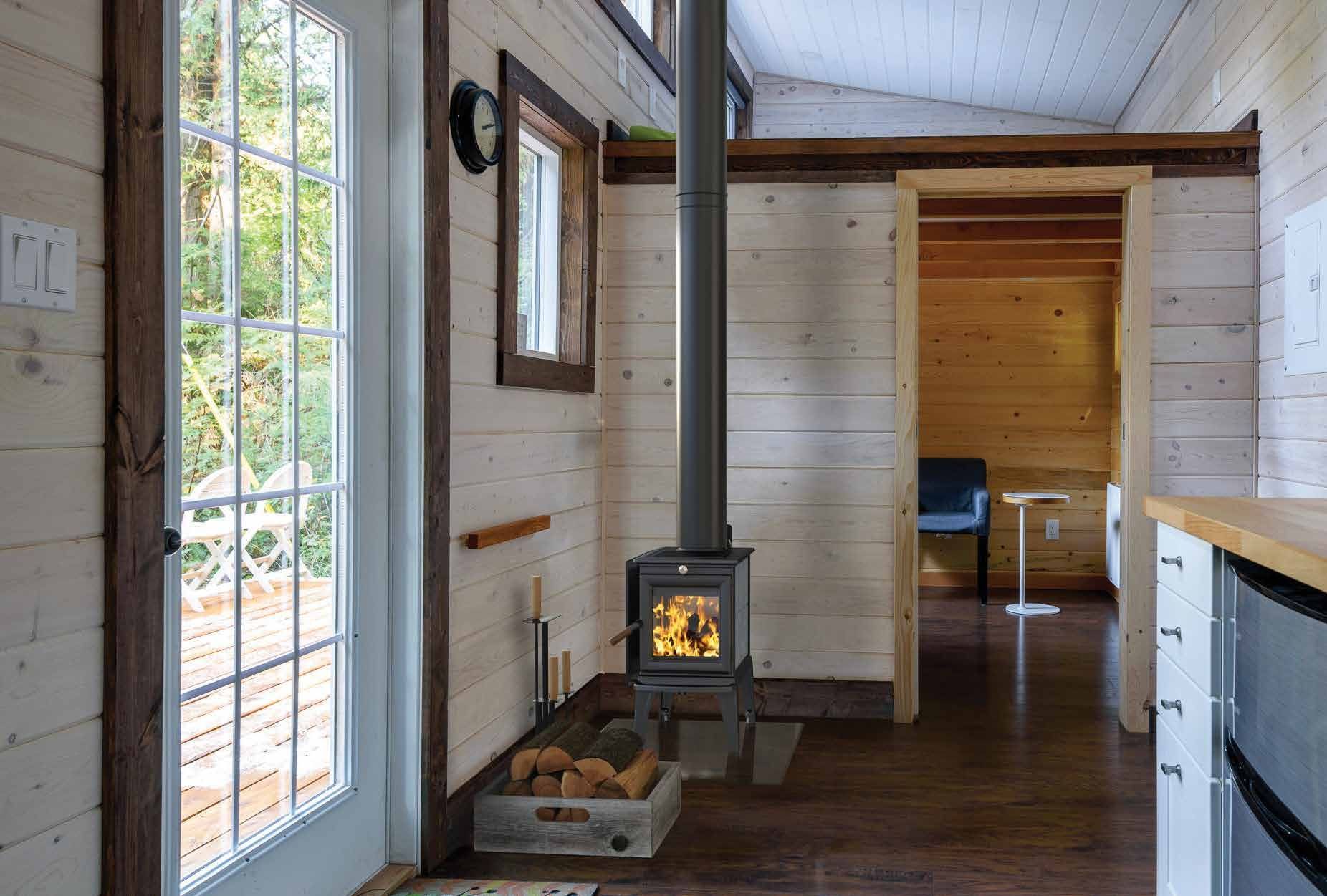 Cozy tiny home interior with a wood-burning stove, firewood, and a view through a glass door. Wooden walls and flooring.