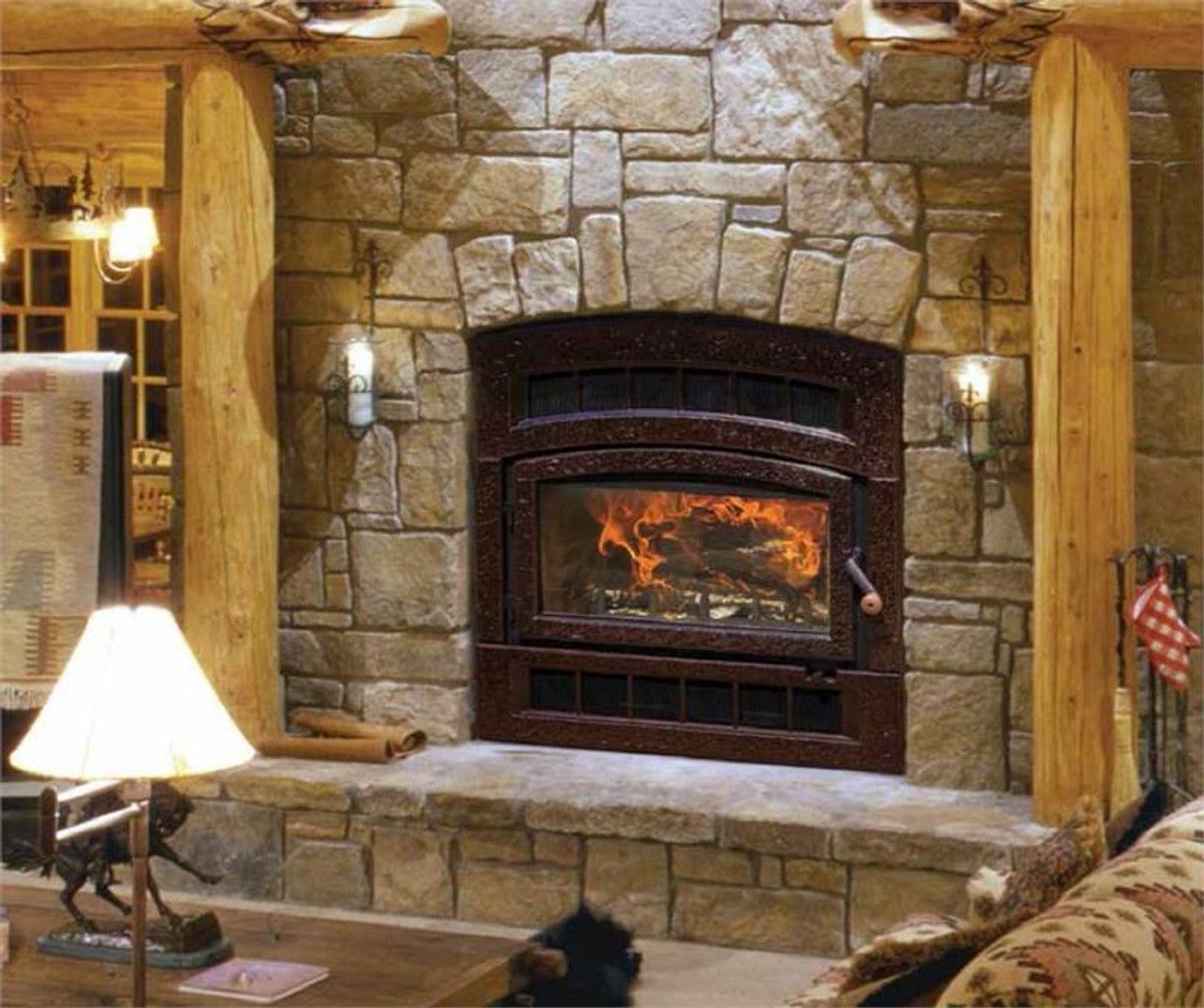 Stone fireplace with a lit fire, built into a stone wall. It's flanked by wooden posts and lit by wall sconces.