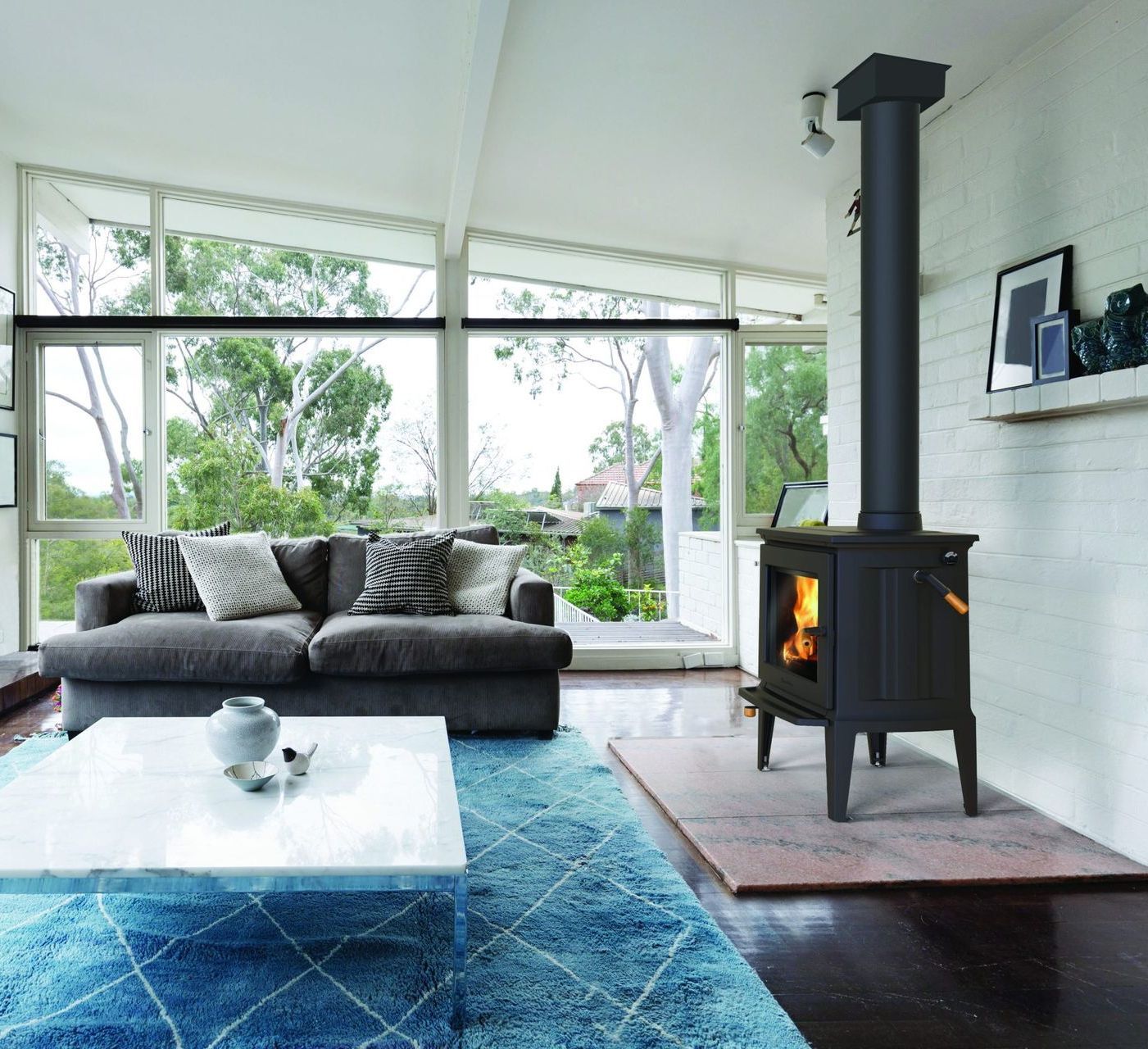 Cozy living room with a fireplace. A dark gray sofa sits in front of large windows overlooking greenery. A blue rug and white coffee table are in the foreground.
