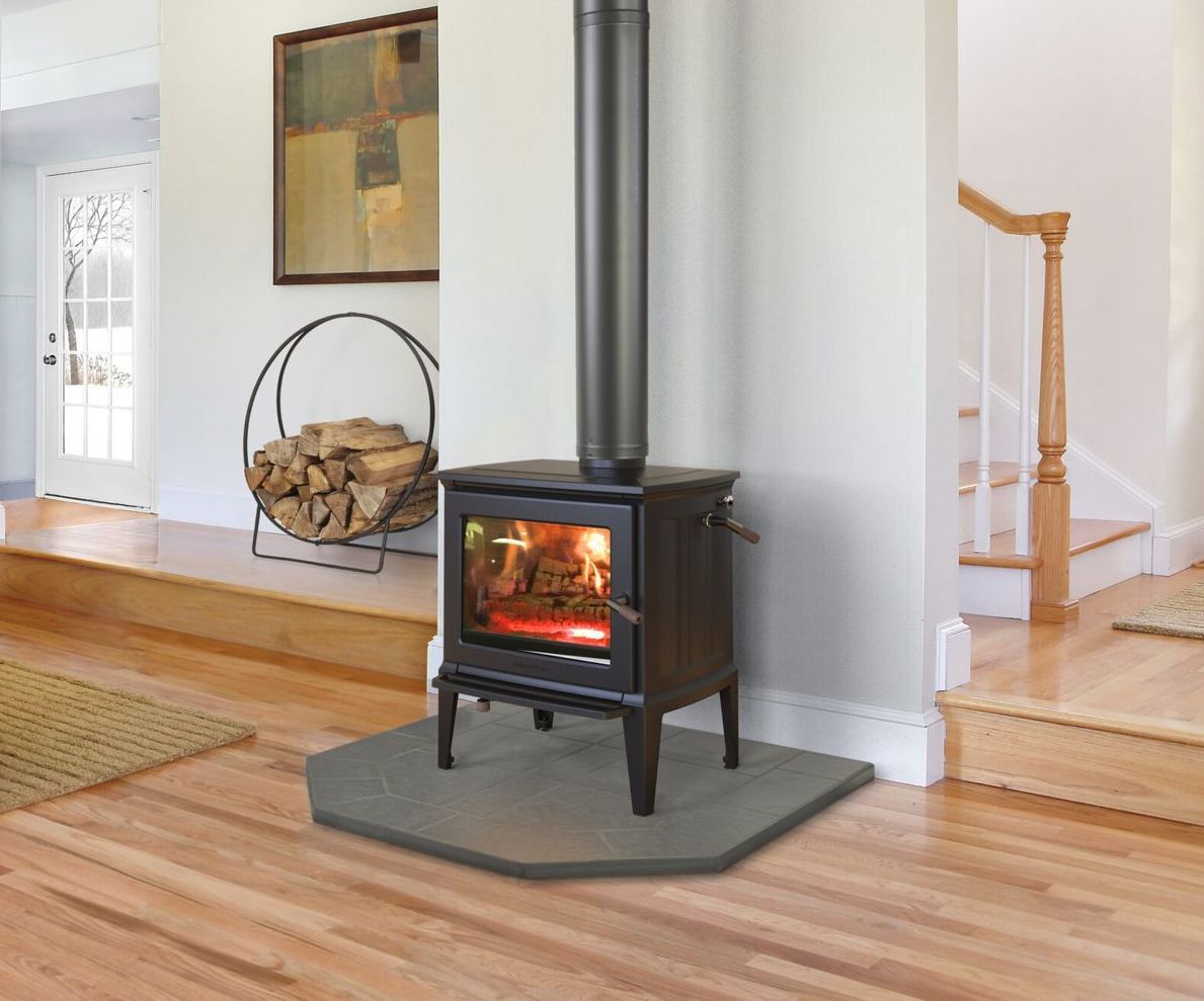 A wood-burning stove with a lit fire sits on a hearth in a home. A decorative firewood rack and stairs are nearby.