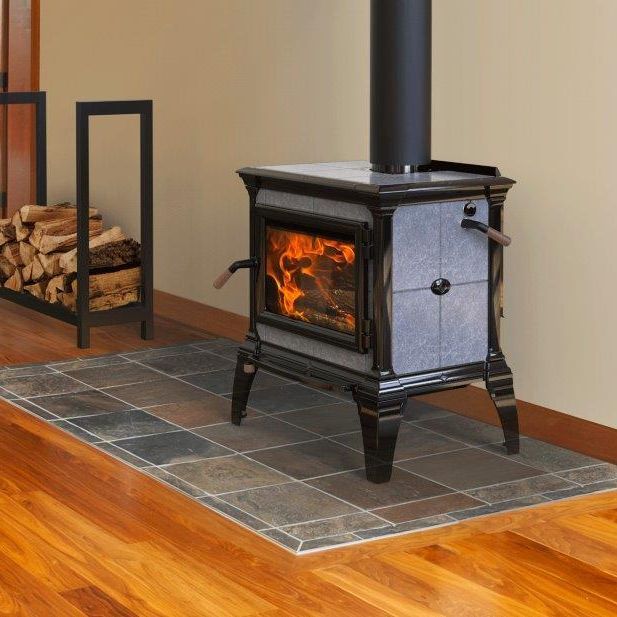 A wood-burning stove with flames sits on a tile hearth. A black firewood holder is to the left.