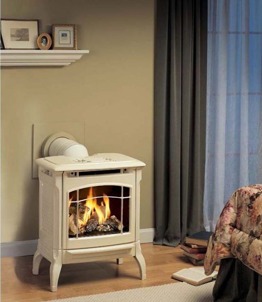 Beige gas fireplace with lit flames in a room. A white vent is attached to the back. A bed, books, and a shelf are visible.