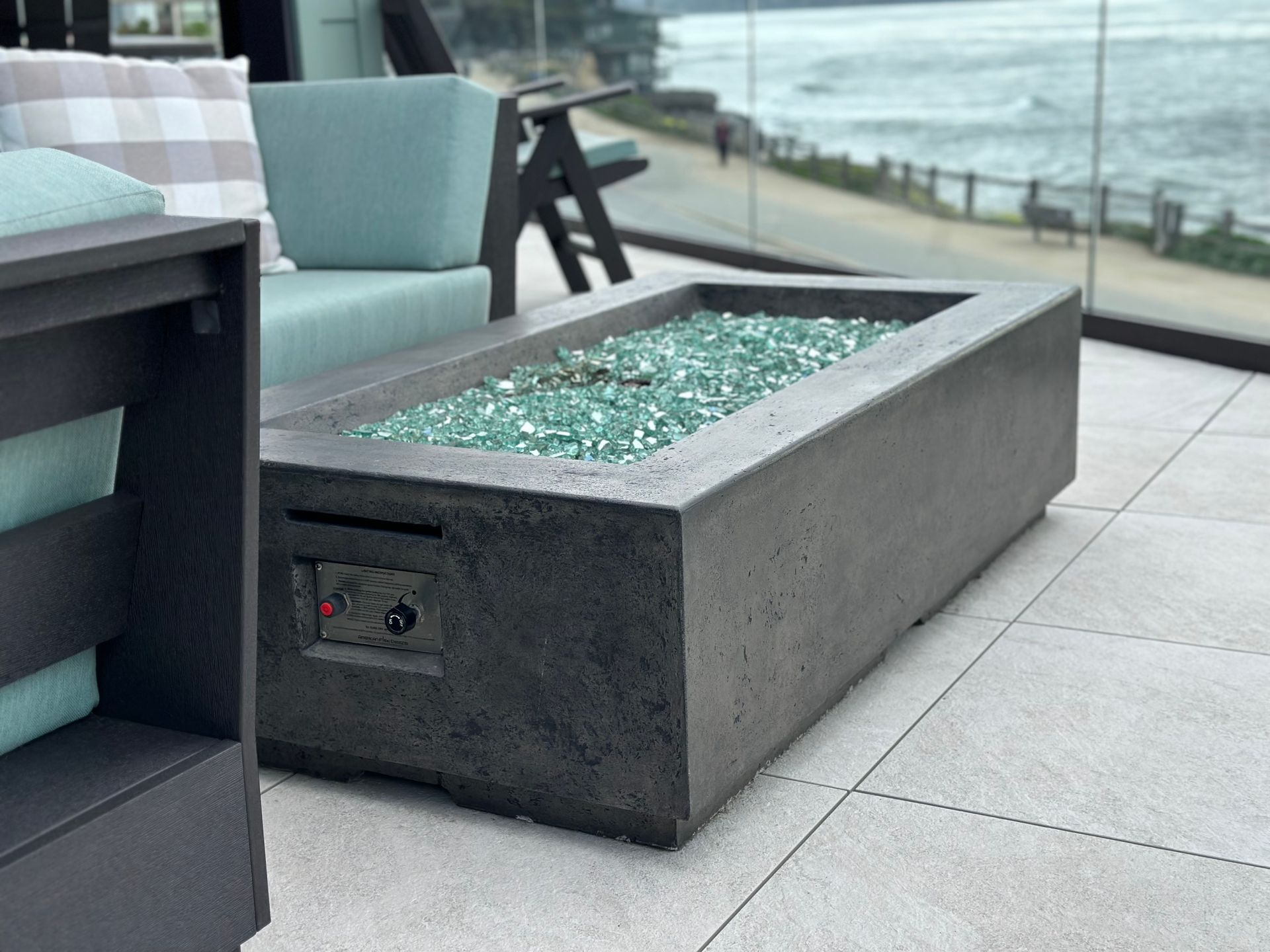 Gray concrete fire pit with turquoise glass stones, next to outdoor seating with ocean view.