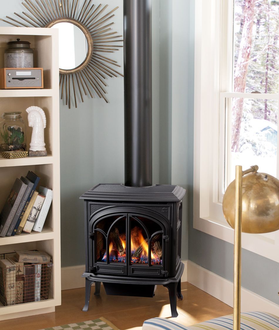 A black gas stove with a lit flame sits in a room with a bookshelf, mirror, and window. A black flue pipe extends upwards.