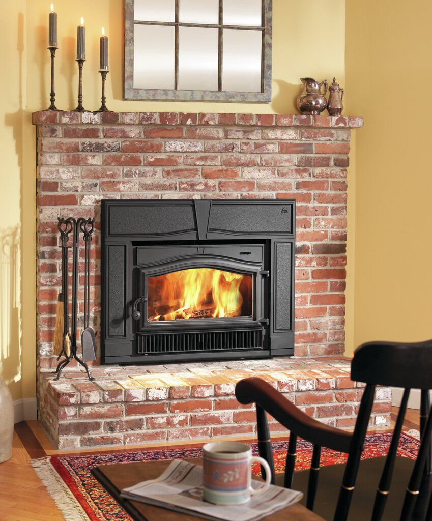 A brick fireplace with a burning fire, topped with a mantle holding candles and a mirror. A chair and mug sit in the foreground.