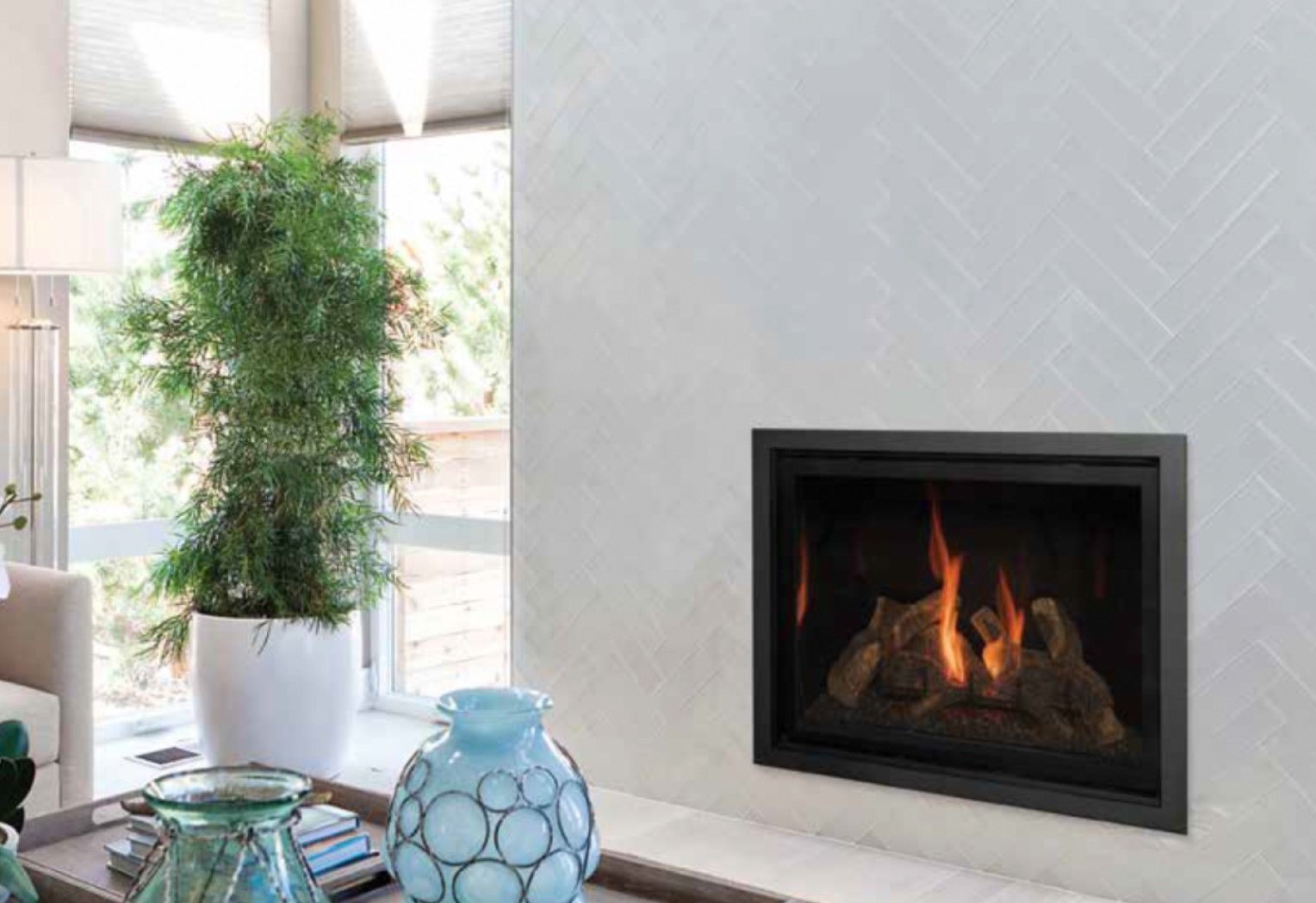 Modern fireplace with flames, set in a white tile wall. A large plant, blue glass vases, and a window are also visible.
