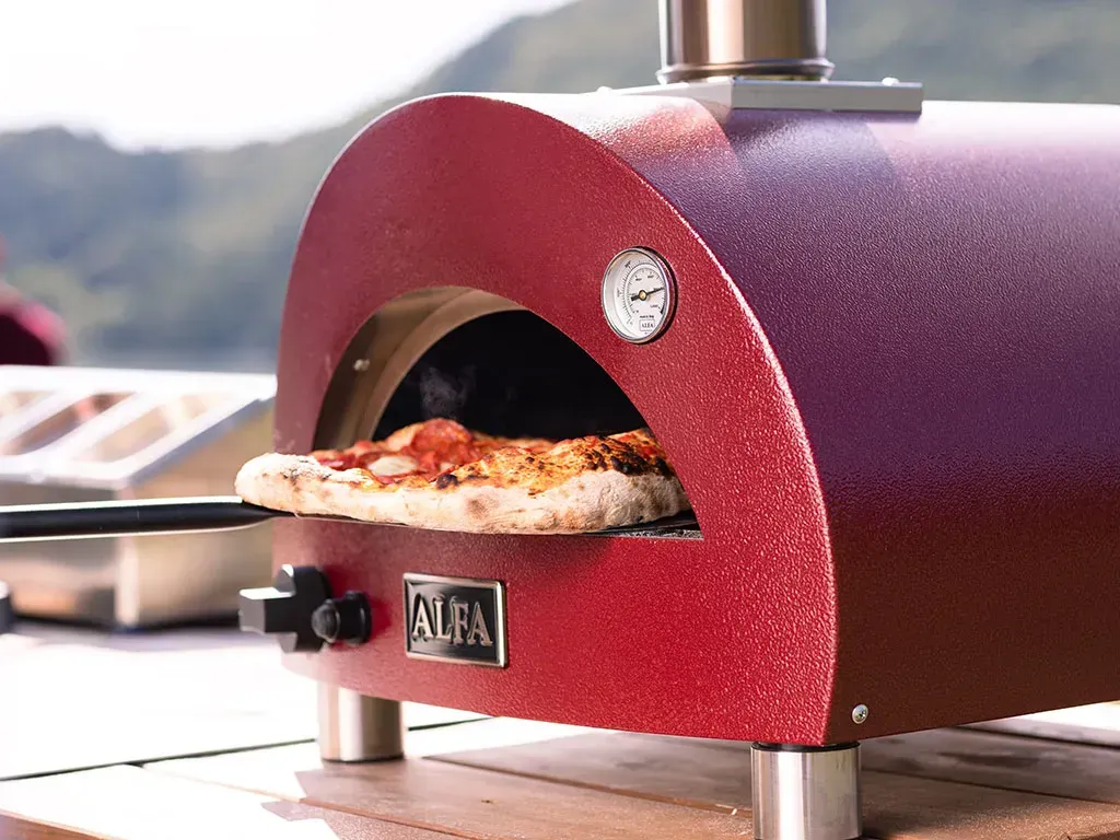 A red outdoor pizza oven with a pizza cooking inside, sitting on a wooden surface.