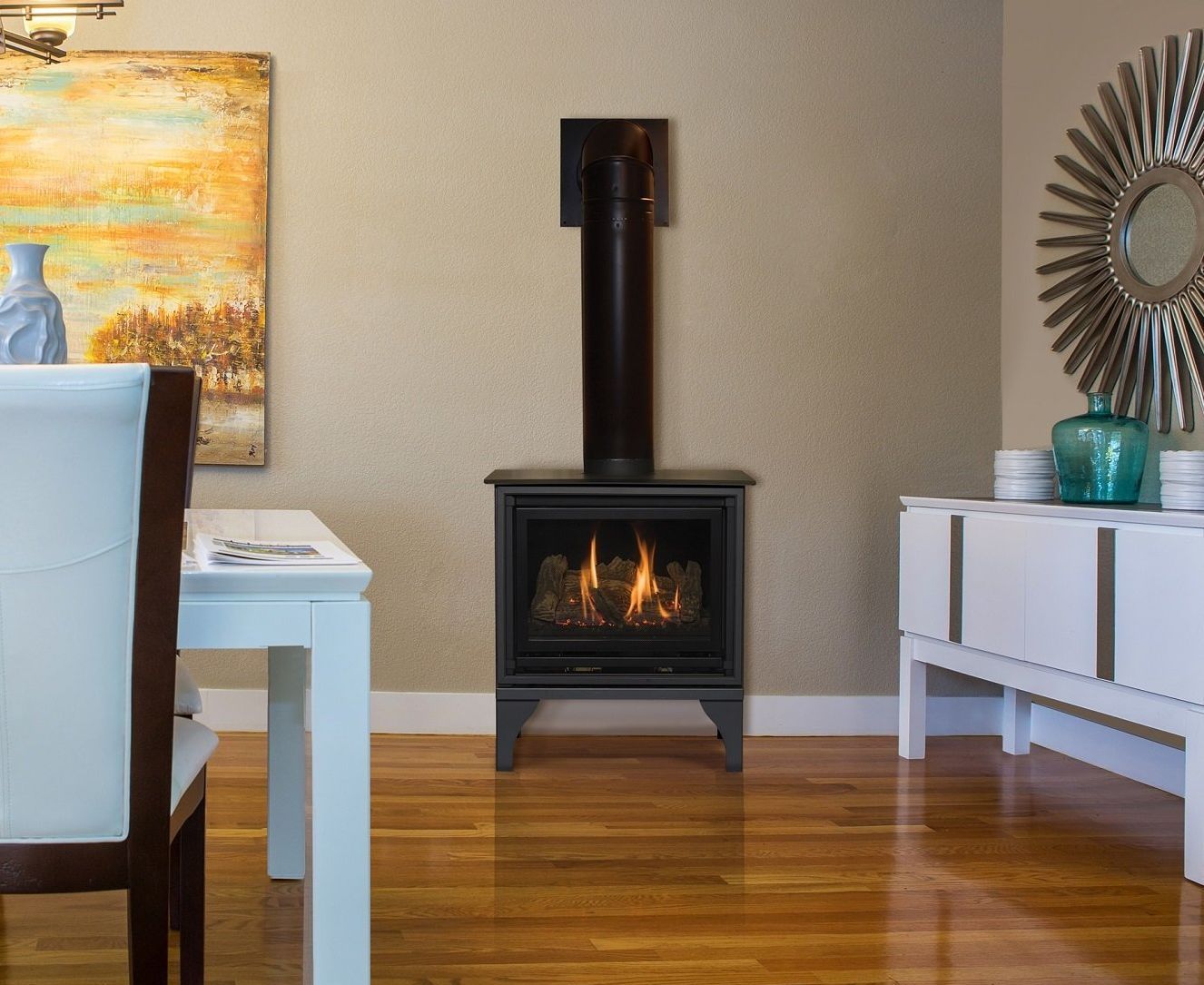 A gas fireplace with a black chimney in a dining room, next to a white buffet and table. A painting hangs on the wall.
