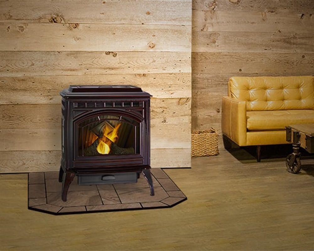 A dark wood pellet stove with a glowing fire sits on a tile hearth in a room with a wood-paneled wall, a yellow couch, and a basket.
