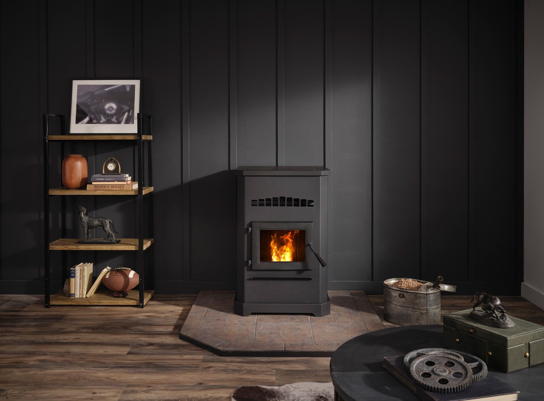 A pellet stove with a fire burning sits against a dark gray paneled wall.  A wooden bookshelf and a dark coffee table are in the room.