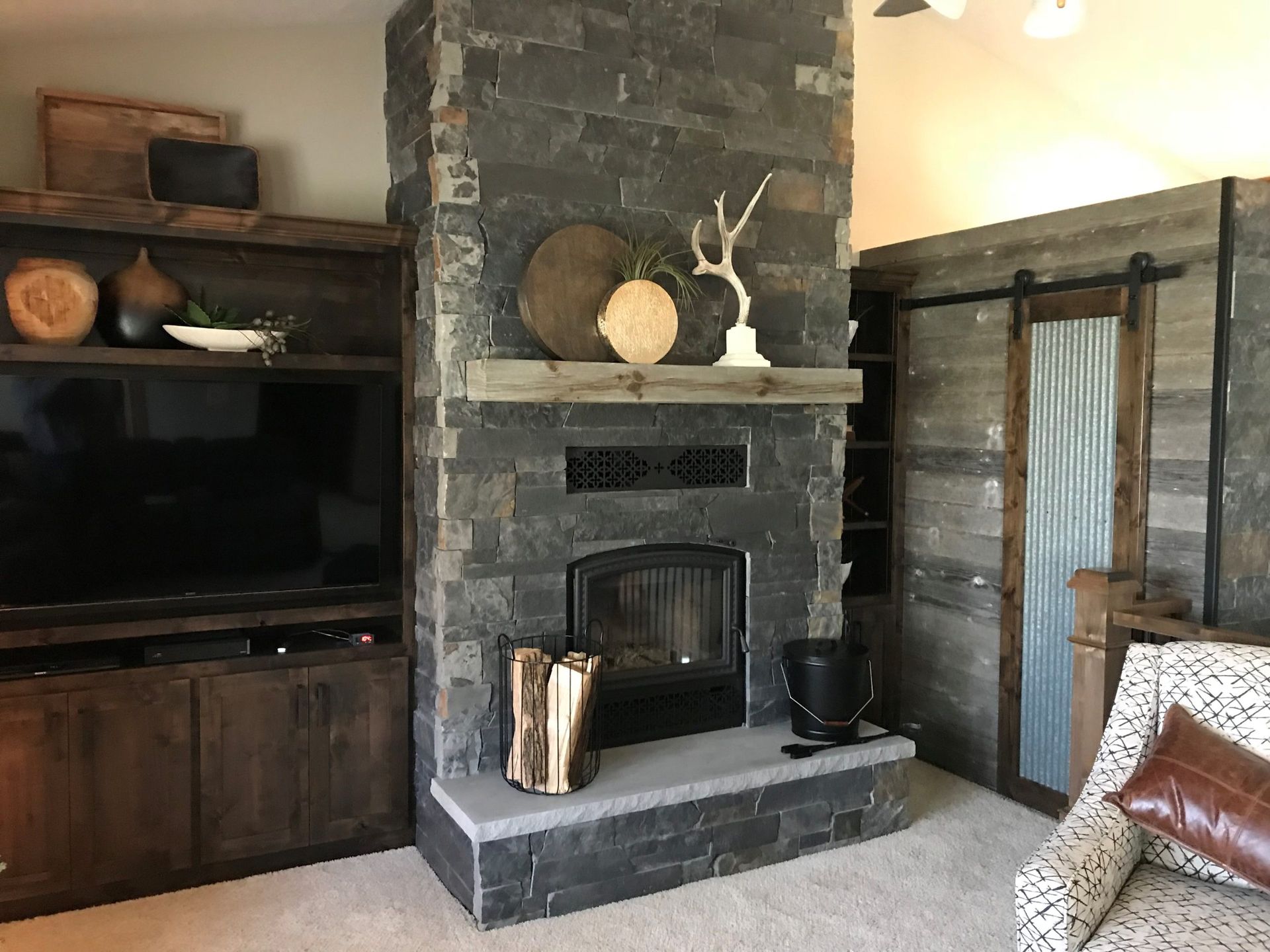 A rustic fireplace with a stone facade, a wooden mantle, and a stack of firewood. A built-in entertainment center and a barn door are also present.