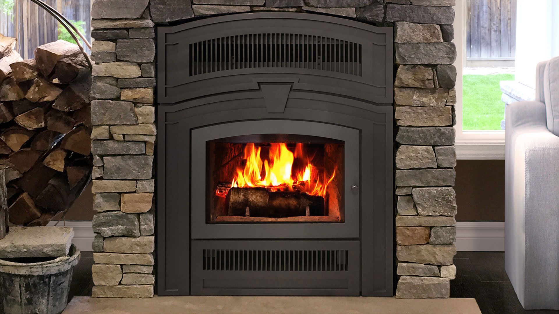 Fireplace with a roaring fire, surrounded by a stone facade. Firewood is stacked to the side.