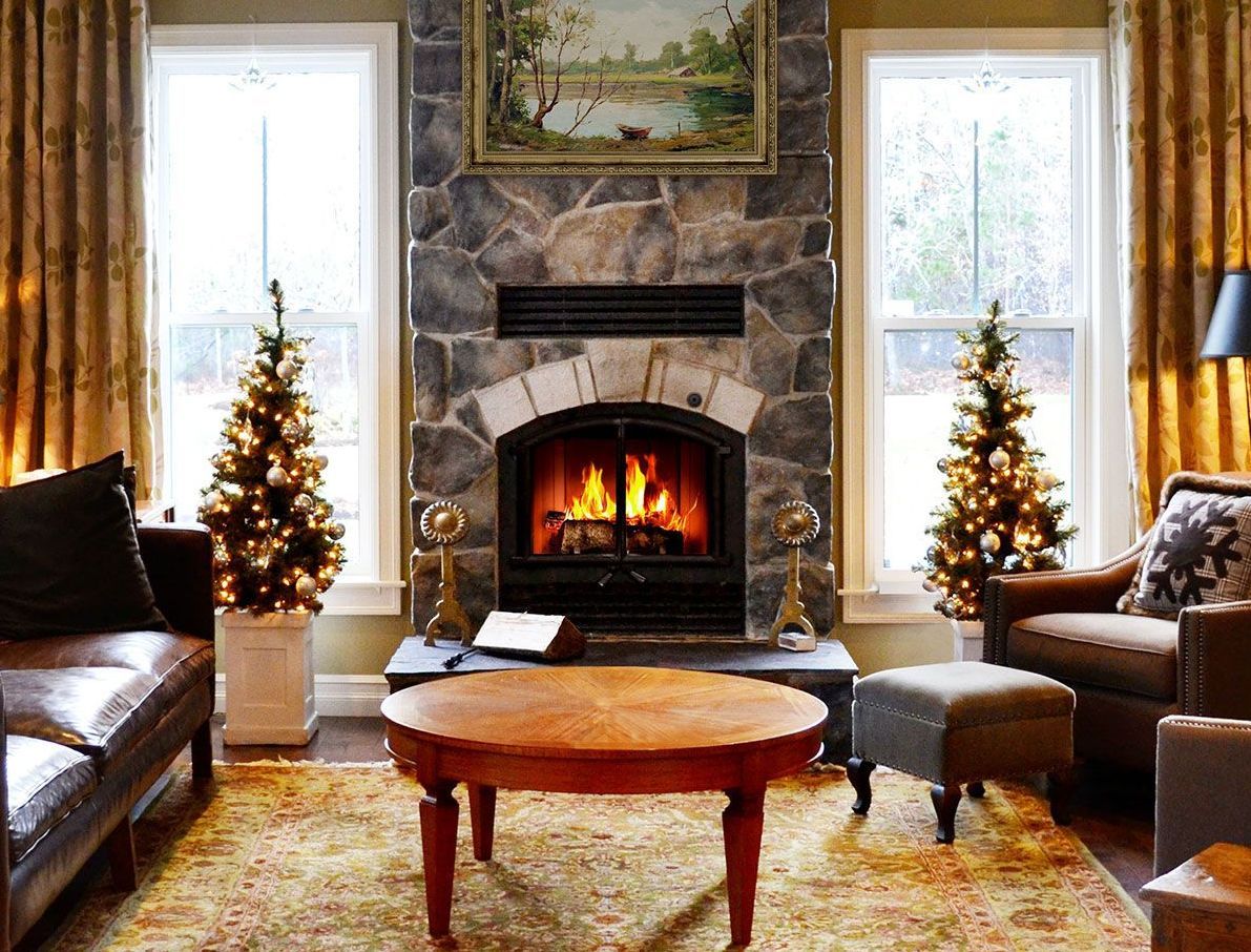 Cozy living room with stone fireplace, flanked by windows and Christmas trees. Brown leather sofa, coffee table, and patterned rug complete the warm setting.