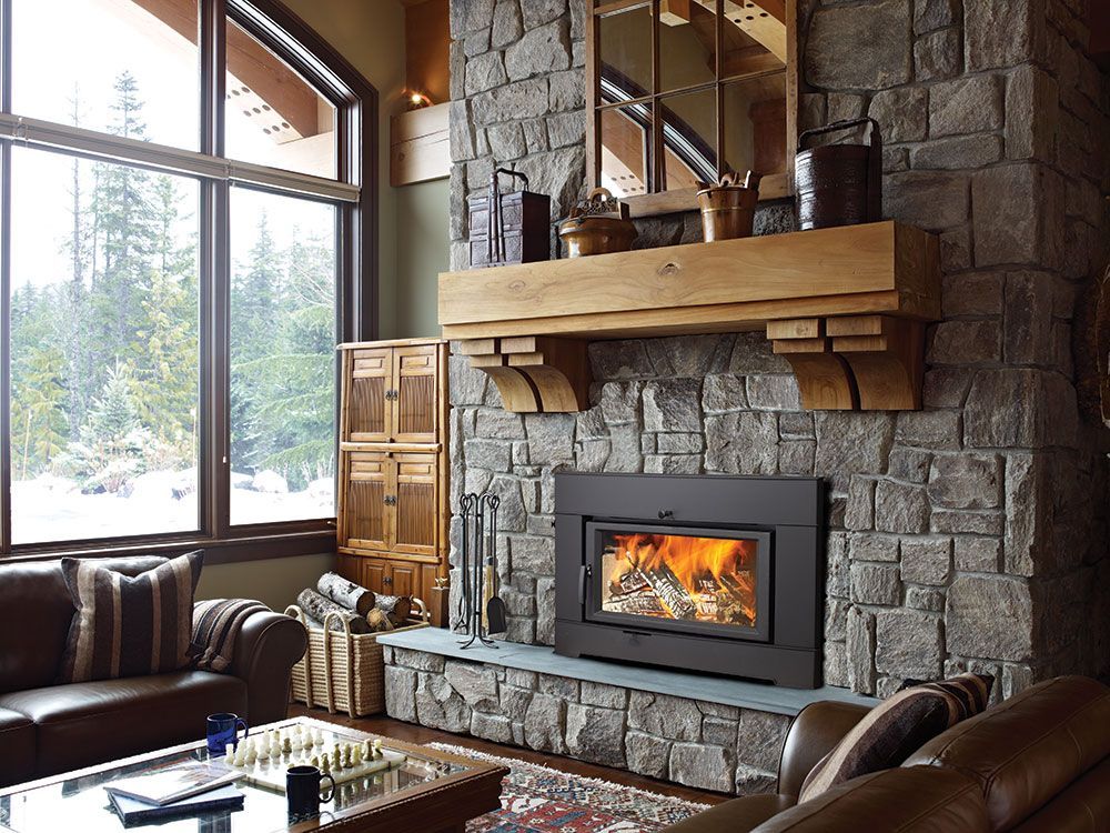 Living room with a stone fireplace, wood mantel, and burning fire. Brown leather couches and a window with a view of trees complete the scene.