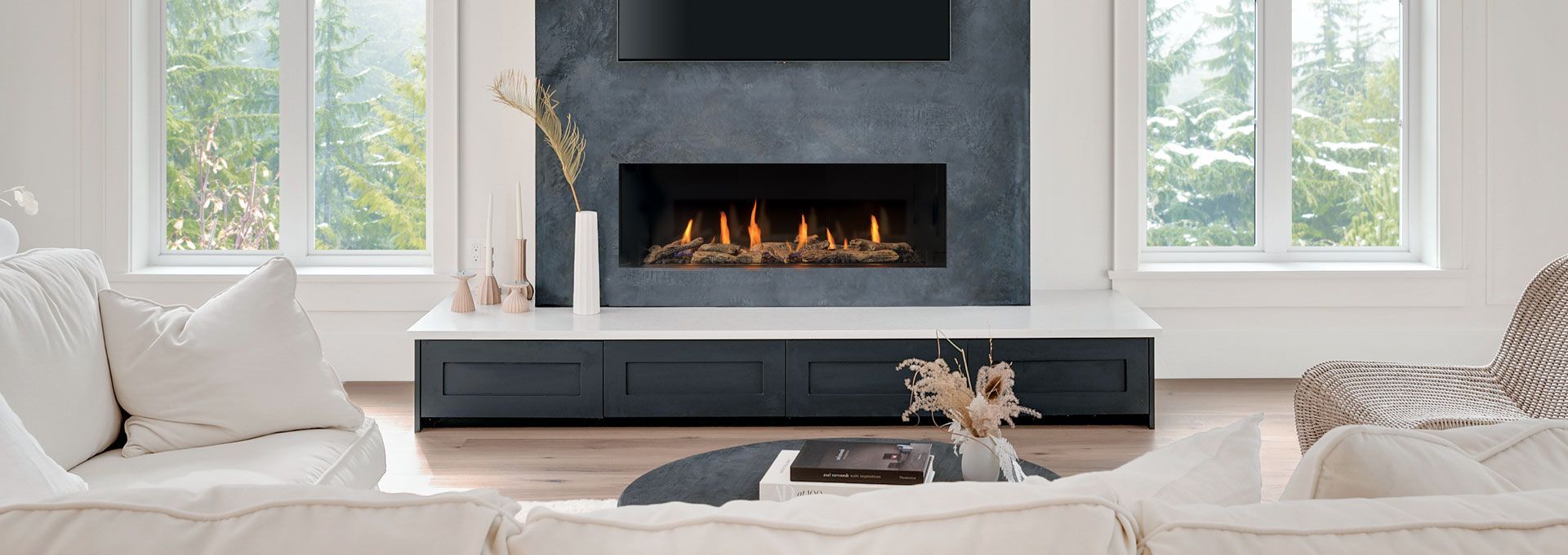 Living room with fireplace, white sofas, and windows overlooking a green forest. Dark blue fireplace, white cabinetry.