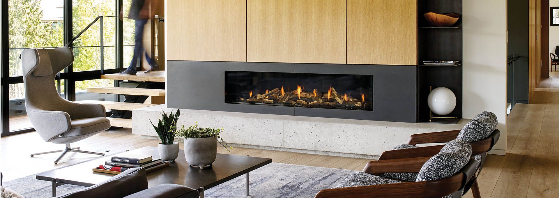 A modern living room with a fireplace, wood paneling, and two armchairs. A person walks up a staircase visible through a window.
